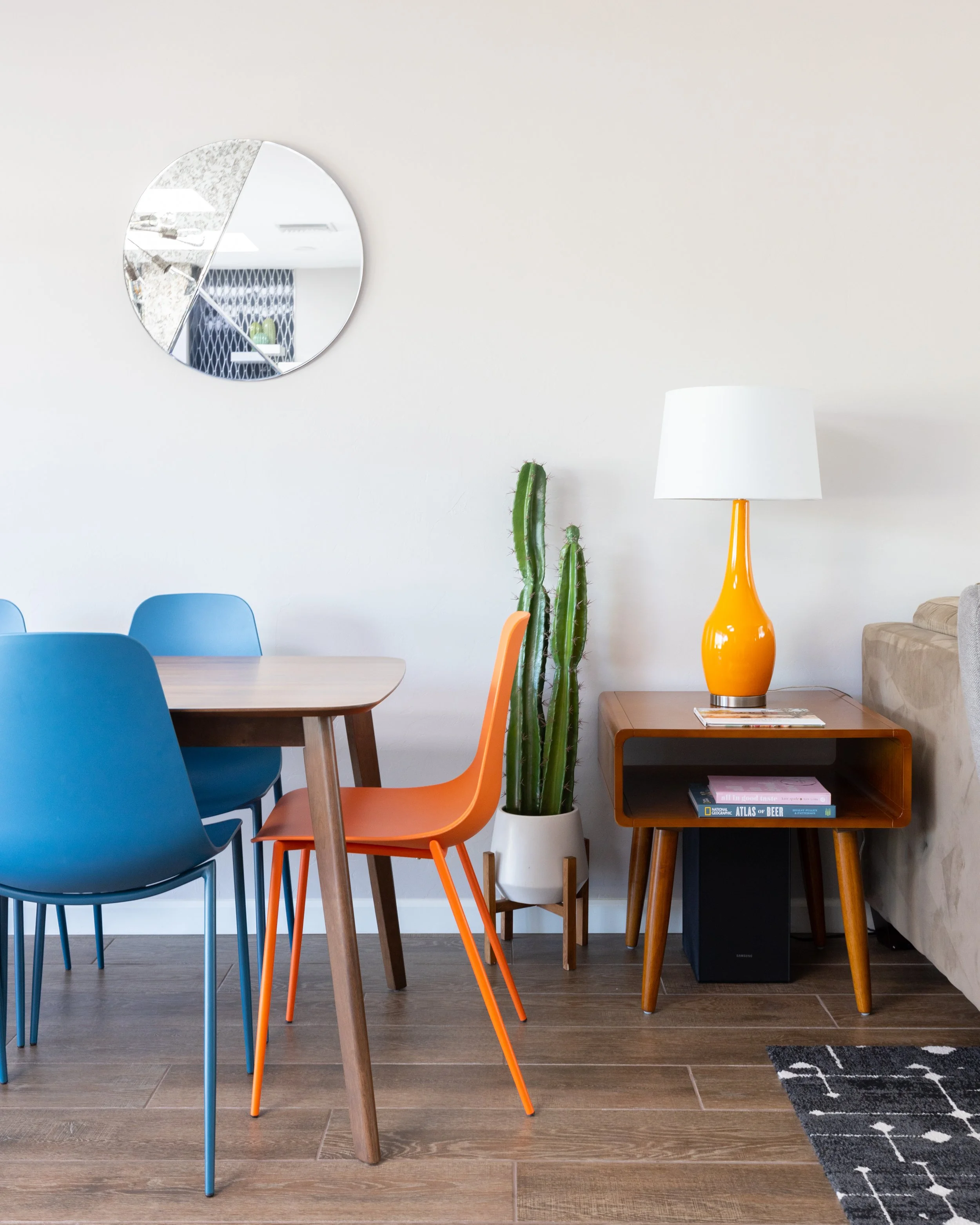Mid-century dining table with blue and orange chairs beside a tall cactus, walnut side table, and bright orange lamp.