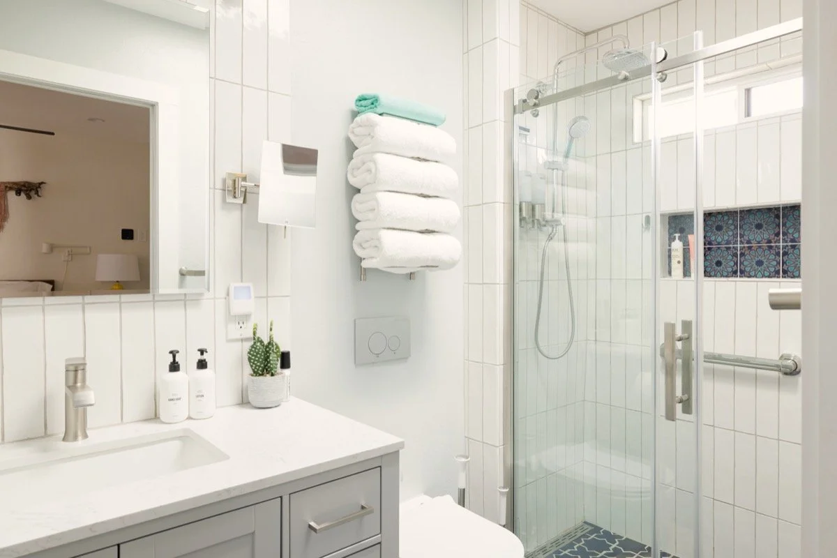 Small white bathroom with a sink vanity, large mirror, stacked towels, wall-mounted toiletries, and a glass-enclosed tiled shower with a blue patterned accent niche.