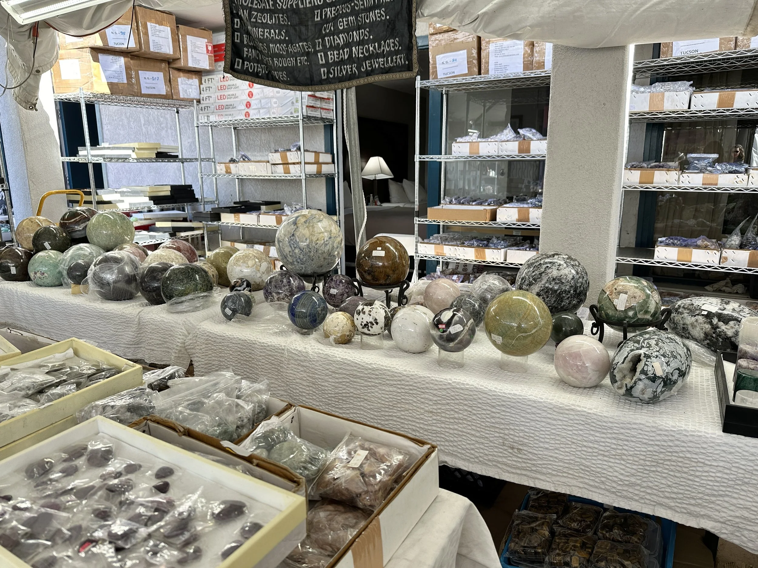 Rows of polished gemstone spheres in green, gray, white, pink, brown, and black displayed on a long table inside a vendor space.