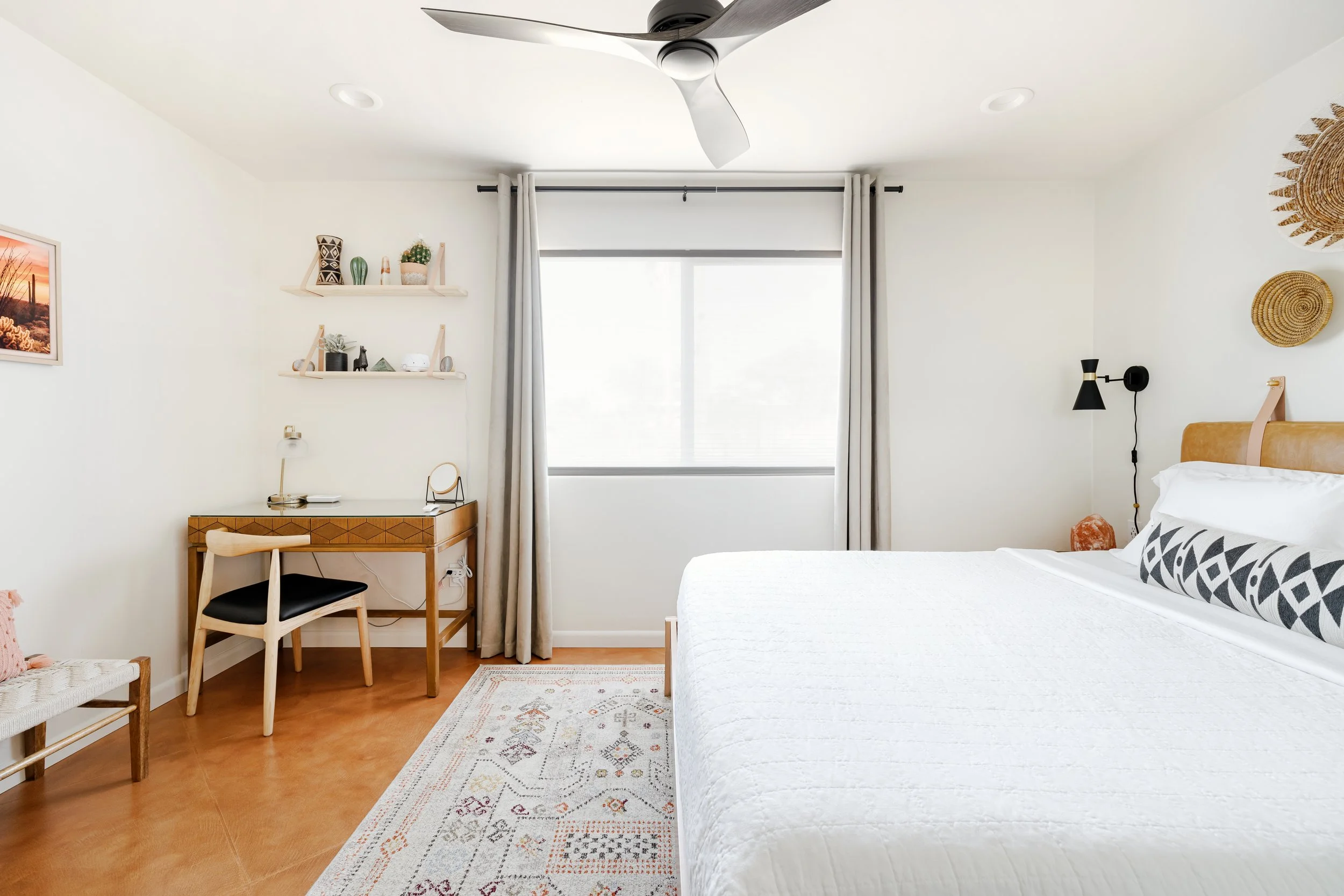 White bed beside a carved wood writing desk, floating shelves, woven wall baskets, patterned rug, and a large curtained window.