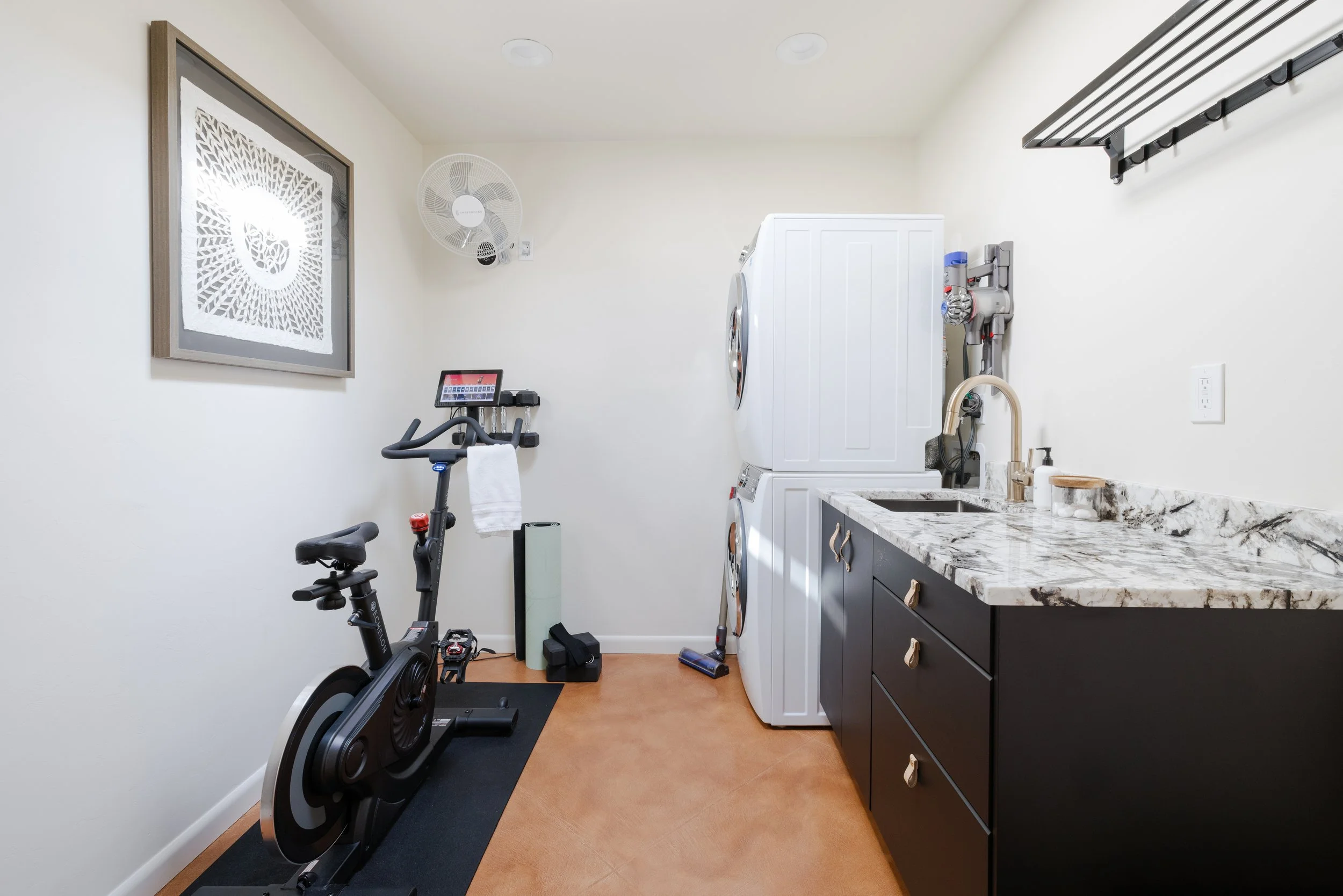 Compact room with a stationary exercise bike, stacked washer and dryer, marble-patterned countertop, dark cabinets, wall fan, and framed artwork.