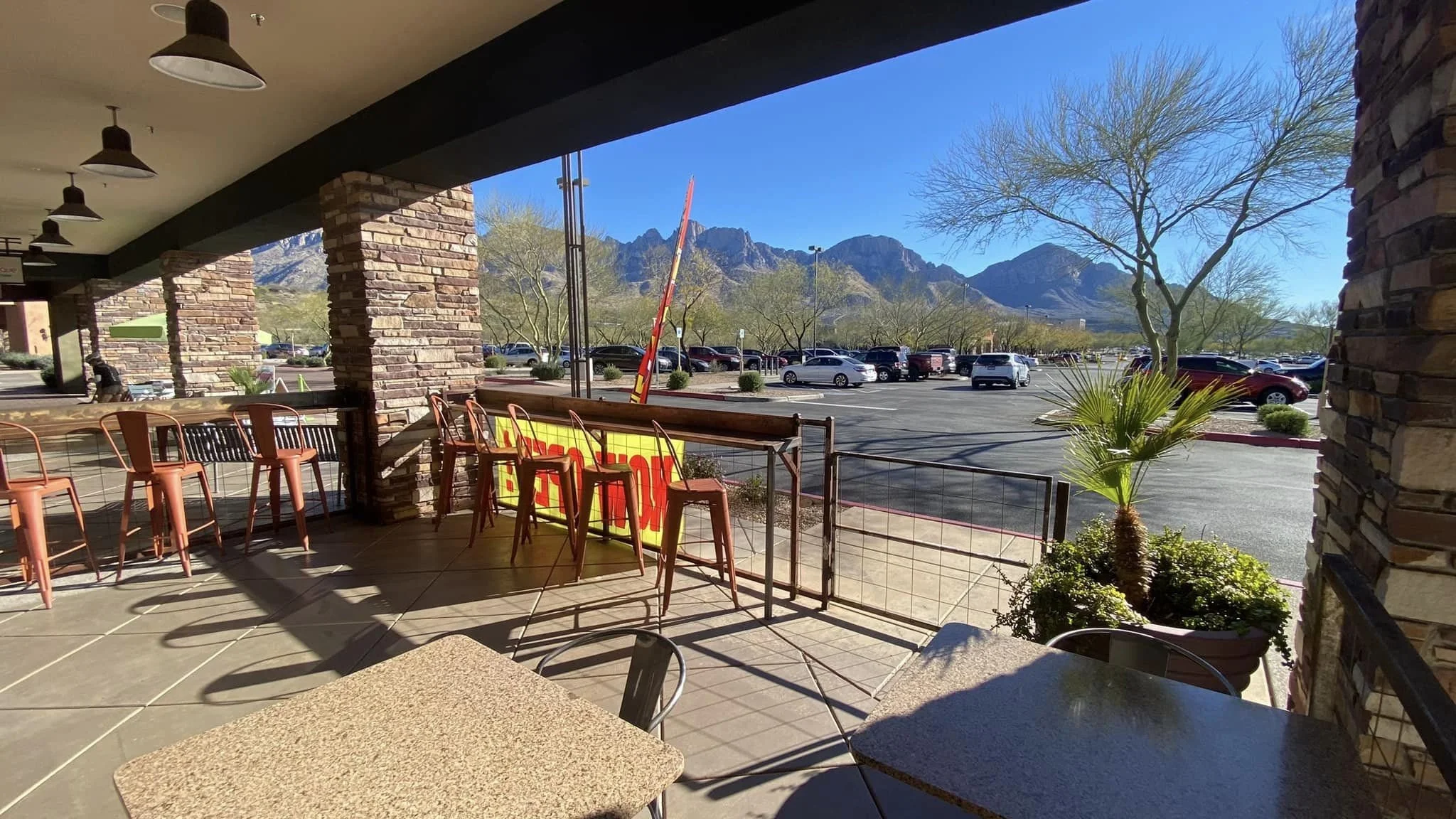 Covered patio at The Hoppy Vine with metal tables and chairs overlooking a parking area and dramatic mountain views beneath a clear blue sky.