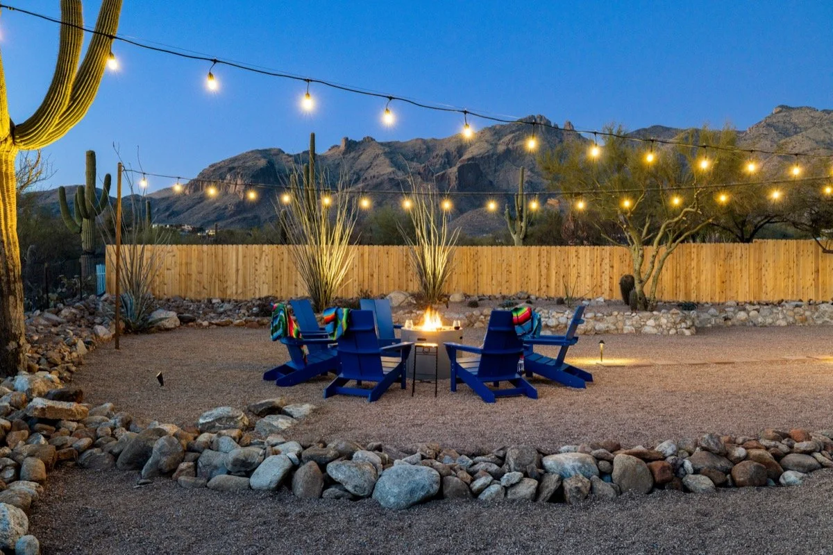 Blue Adirondack chairs circle a glowing fire pit beneath string lights, with saguaros and rugged Catalina Mountains beyond the yard.