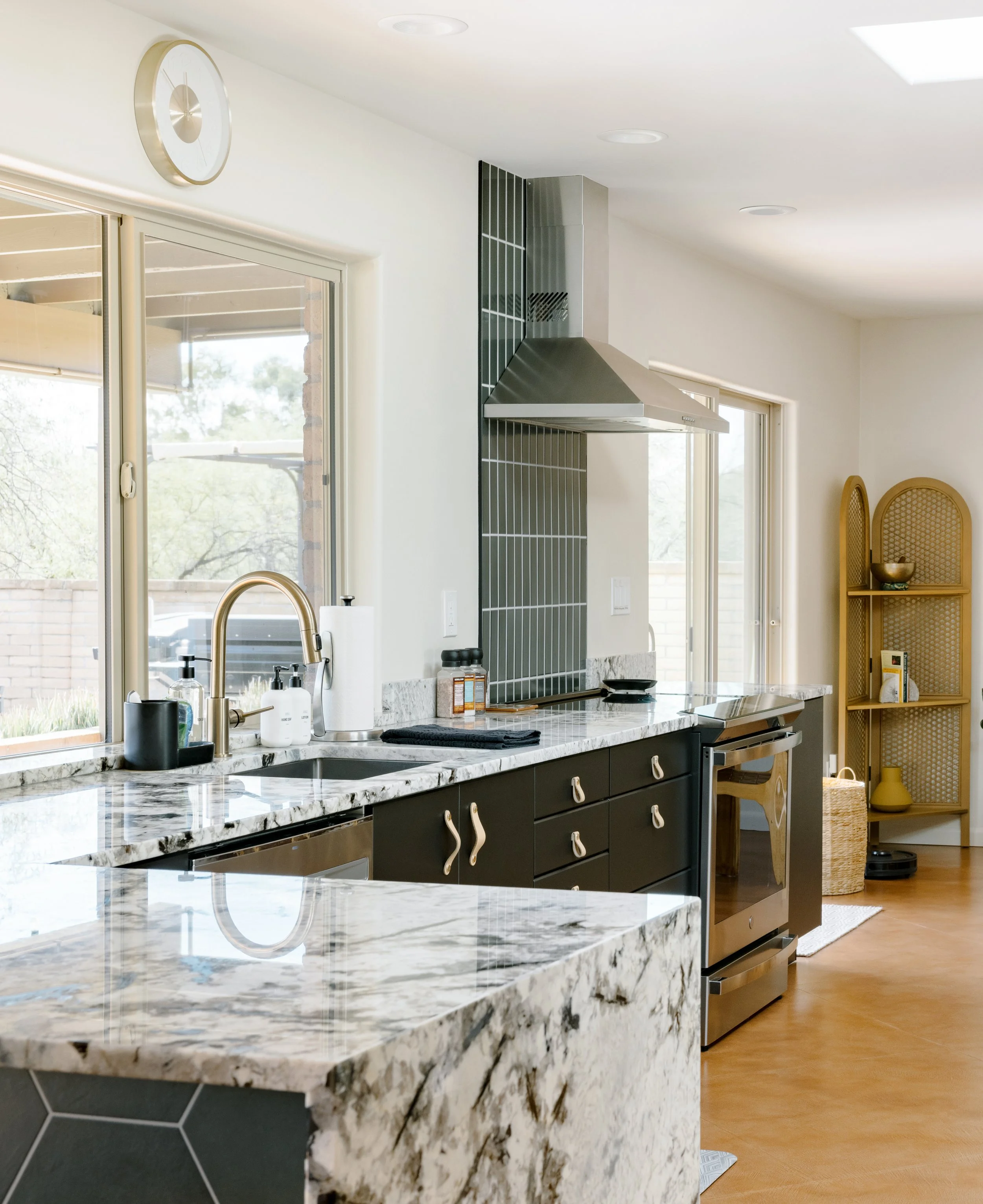 Kitchen with a large marble-patterned island, black cabinetry, stainless steel range hood, and windows above the countertops.