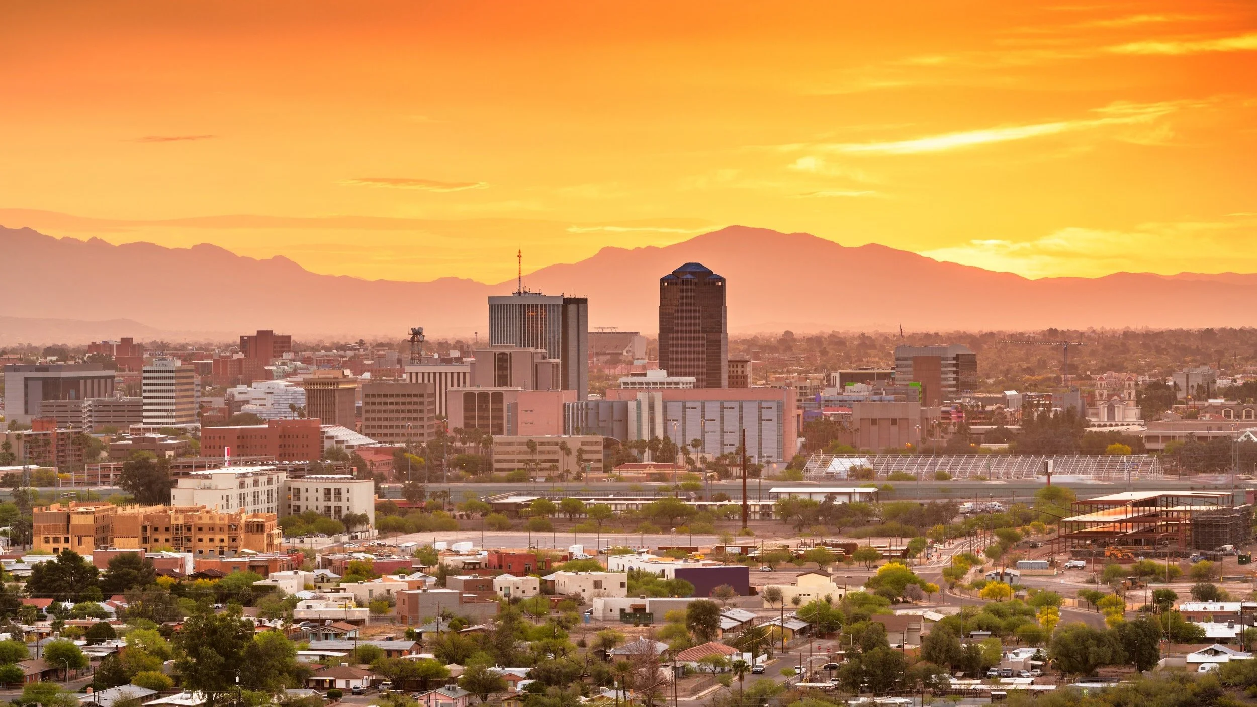City skyline spreads across the foreground beneath a glowing golden-orange sky, with layered mountains silhouetted in the distance.