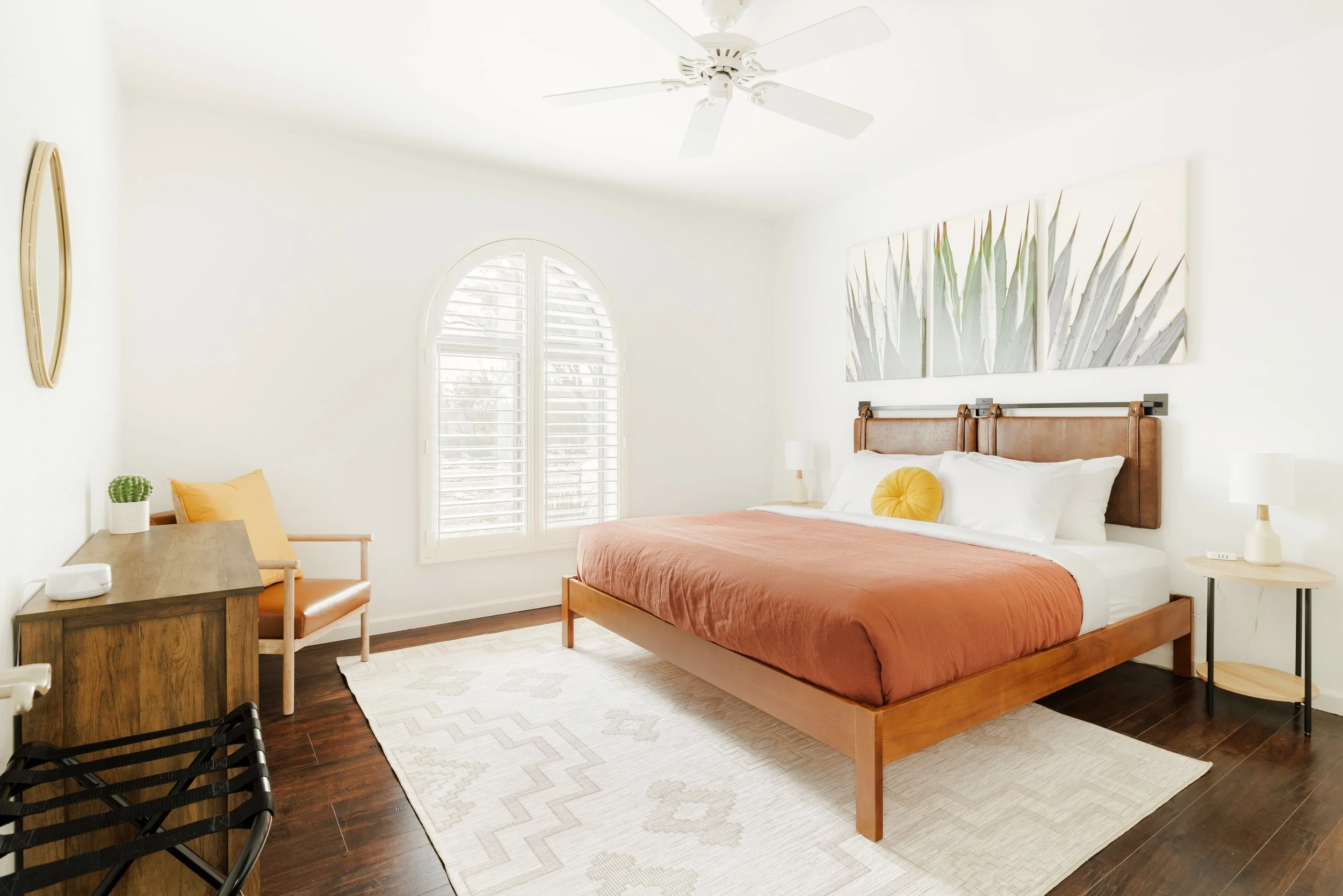 Bright bedroom with rust-colored bedding, leather headboard, large agave artwork, arched window, wood desk, and mustard accent chair.