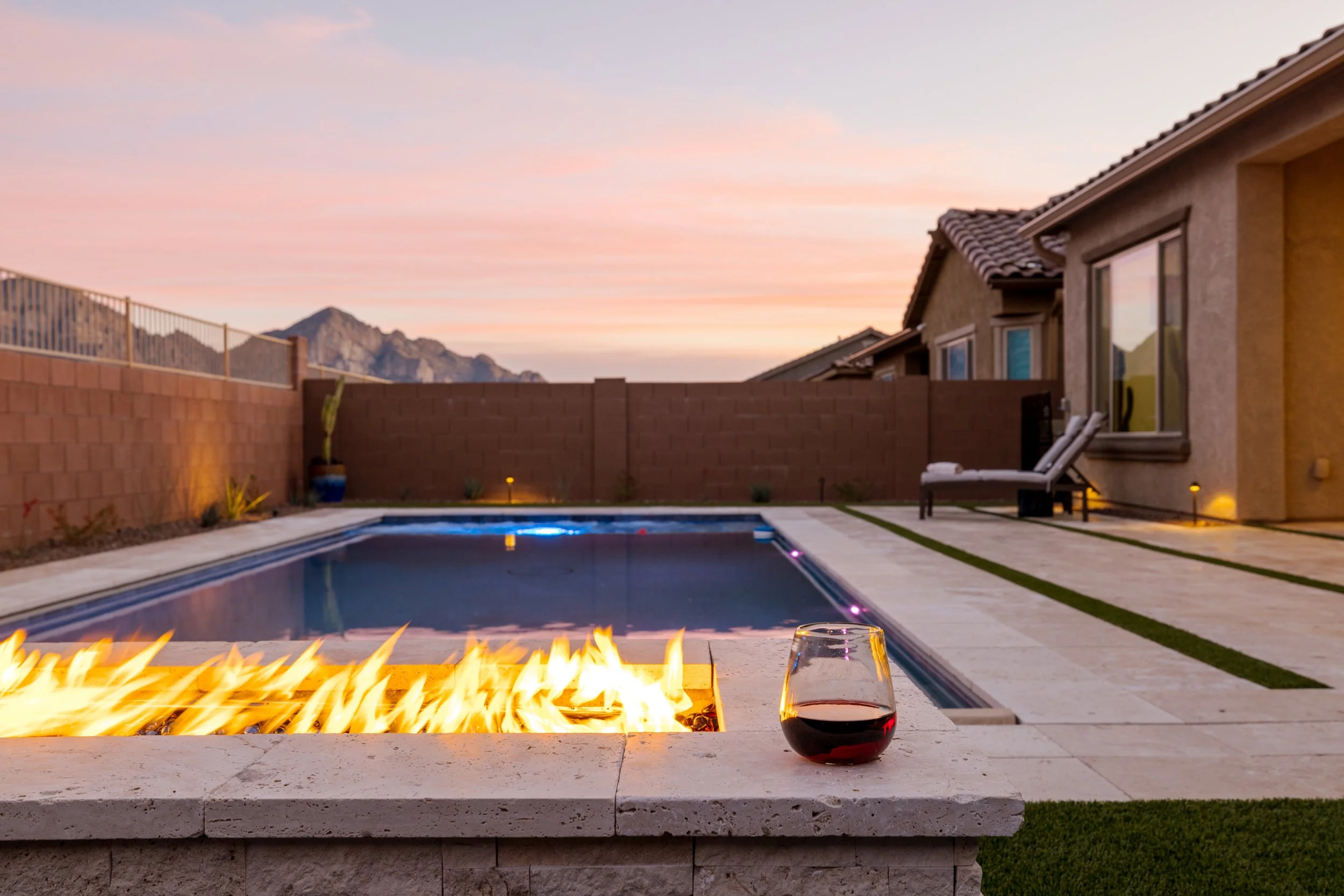 Backyard pool at Casa Alpenglow at sunset with a long fire feature burning beside the water, a glass of red wine on the ledge, lounge chairs, and mountain views in the distance.
