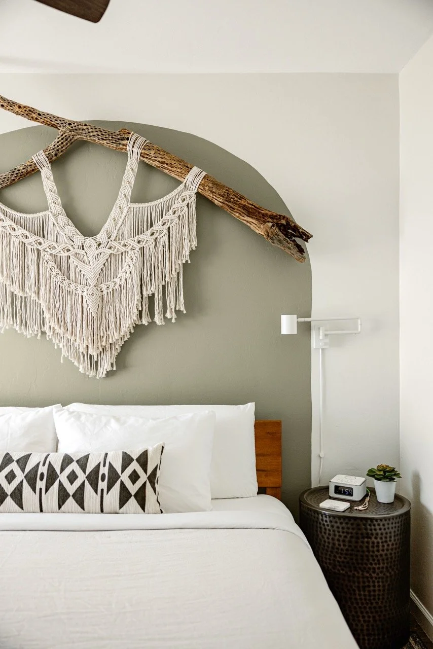 A white bed with geometric patterned pillow in front of an olive green accent arch. A white macramé piece on a cholla branch hangs behind, with a metal barrel side table and a white mounted light to the right.