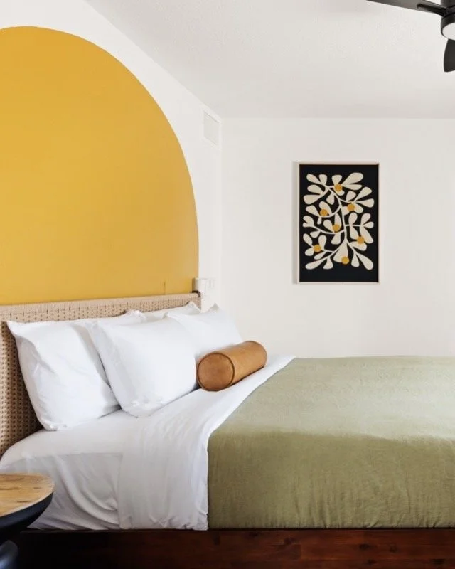 King bedroom with an arched mustard accent wall, framed artwork, white bedding, and soft natural light.