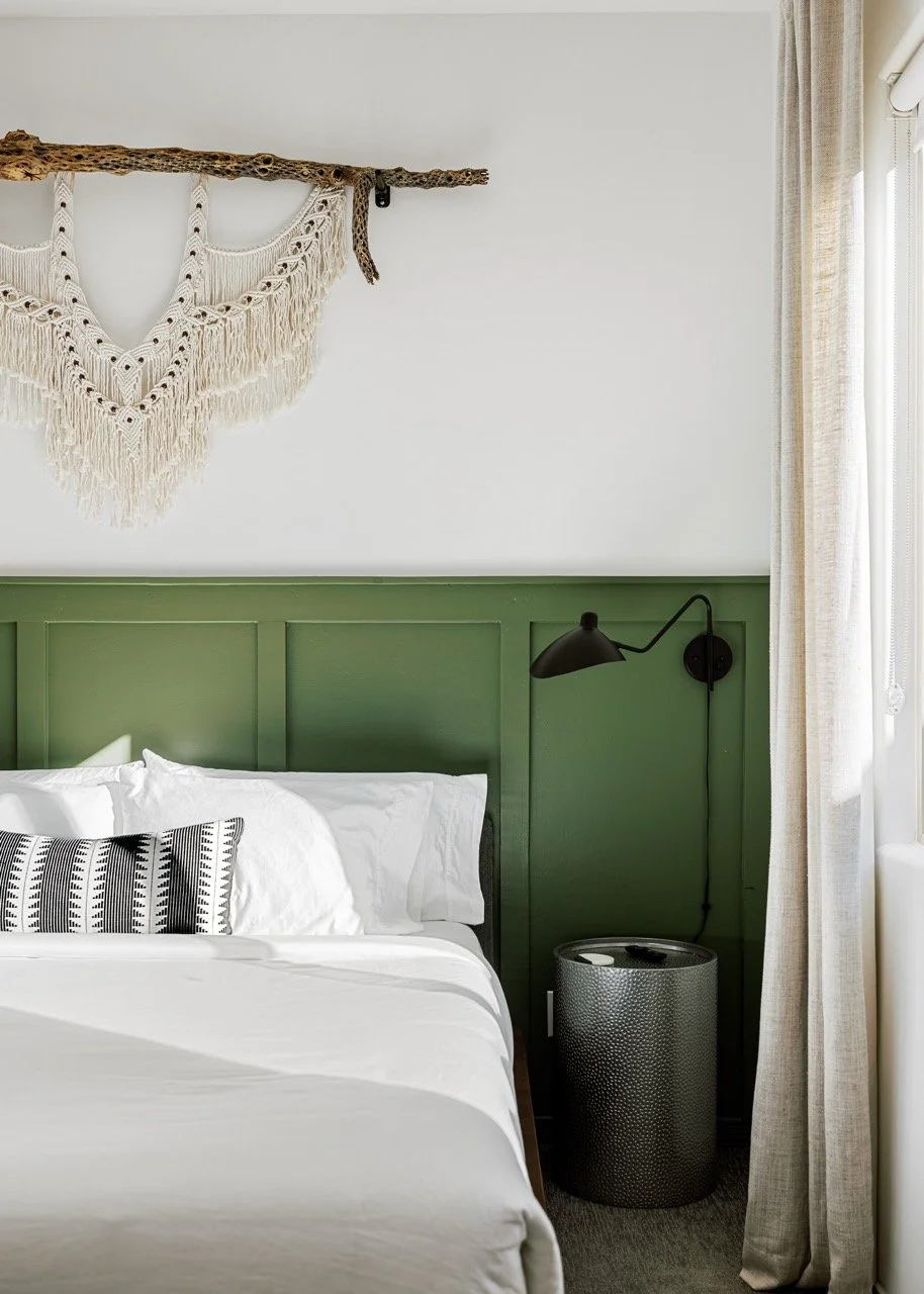 A natural branch with a cream macramé piece hangs above deep green wainscoting. White bedding with patterned accent pillows, a black wall-mounted reading lamp, metal nightstand, and soft linen curtains complete the room.