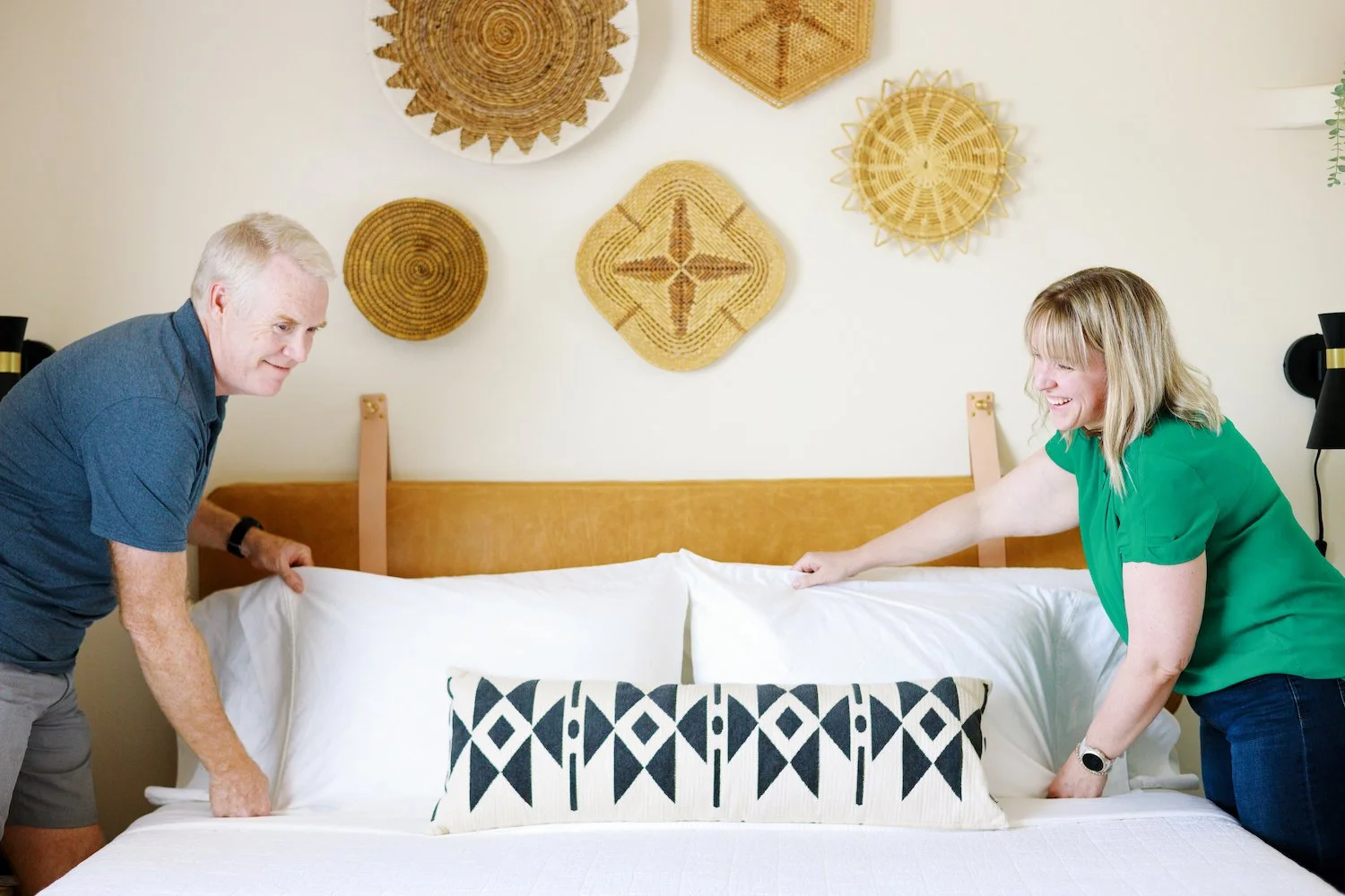 Blonde woman and gray-haired man smooth white pillows onto a neatly made bed beneath woven basket wall art.