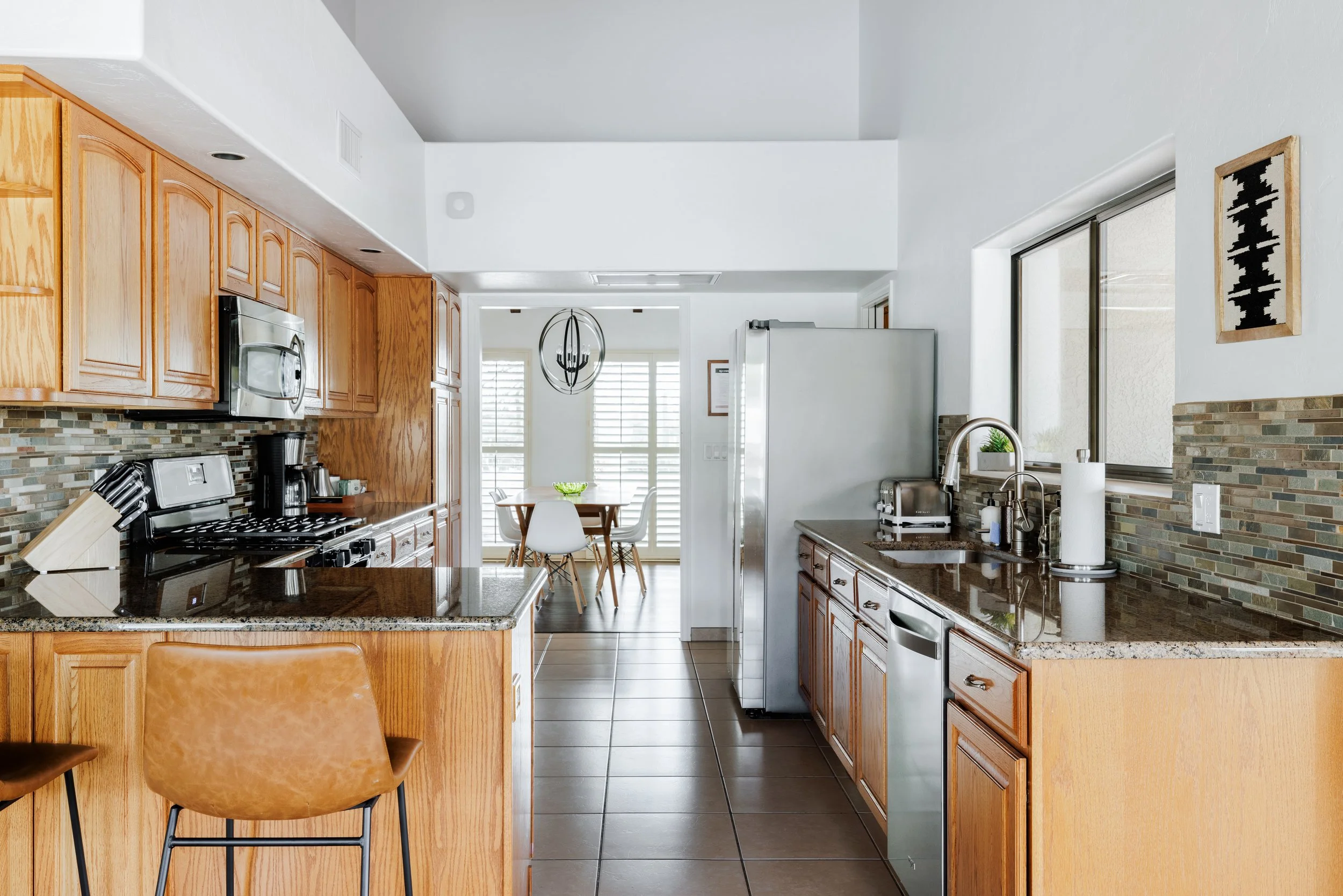 Kitchen with bright, natural light, wood cabinetry, granite countertops, a breakfast bar, stainless steel appliances, tile flooring, and a view of the dining room in the background.