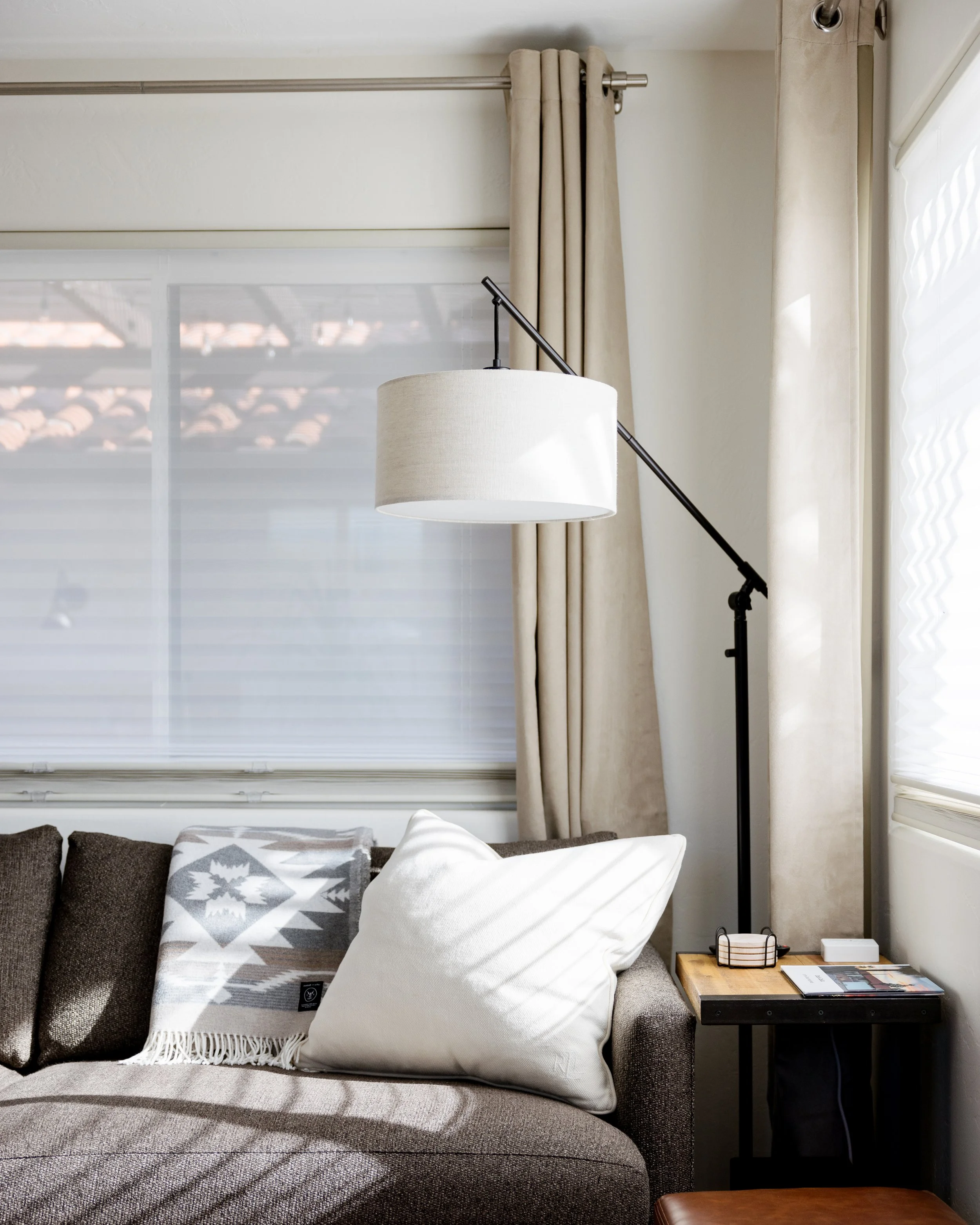 Dark gray sofa beneath windows with cream curtains. A black floor lamp, side table, and patterned throw pillows create a bright, comfortable seating area.