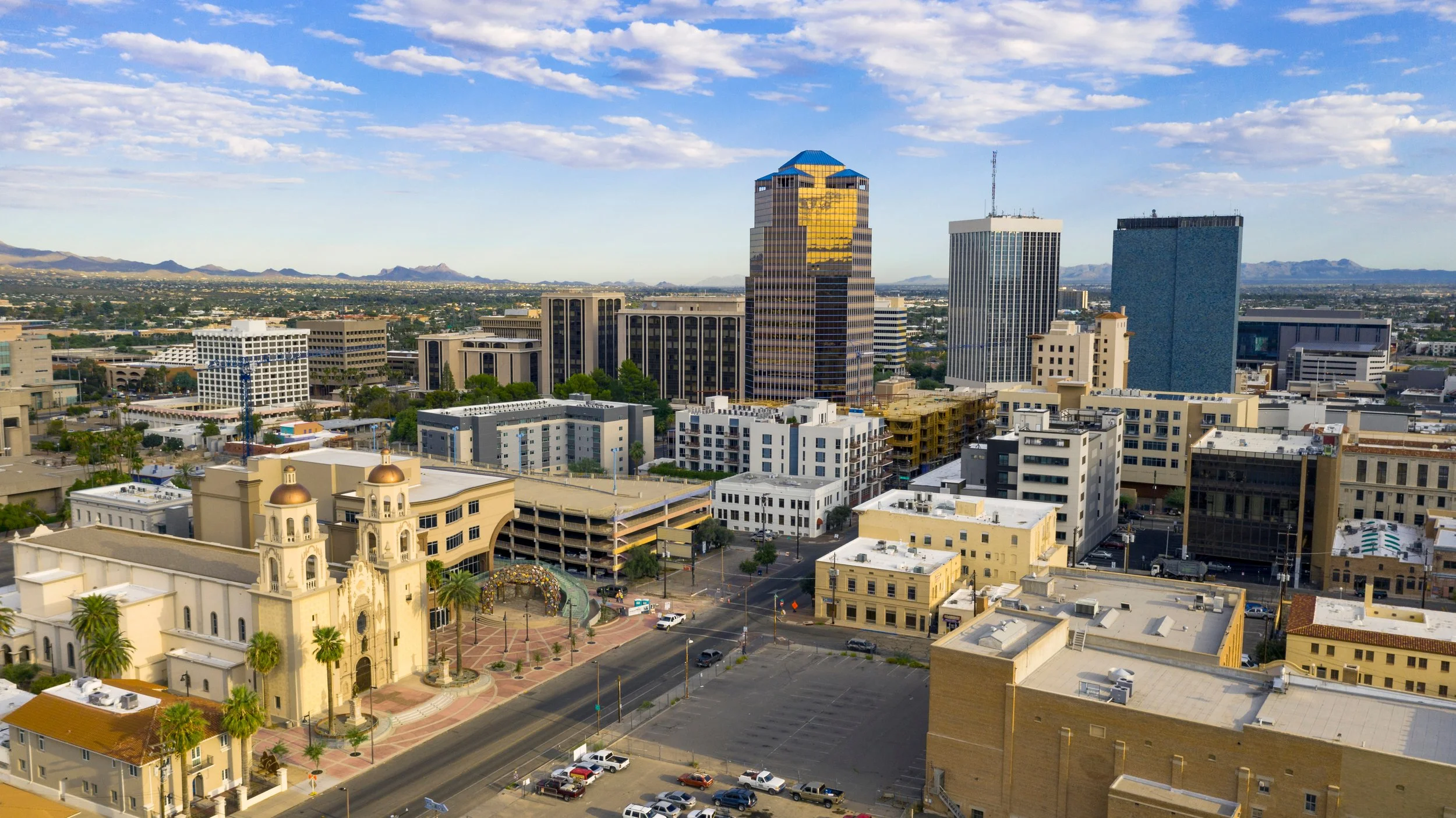 Elevated view of a city center with office towers, low-rise buildings, streets, and a large cream-colored church with two domed towers in the foreground.