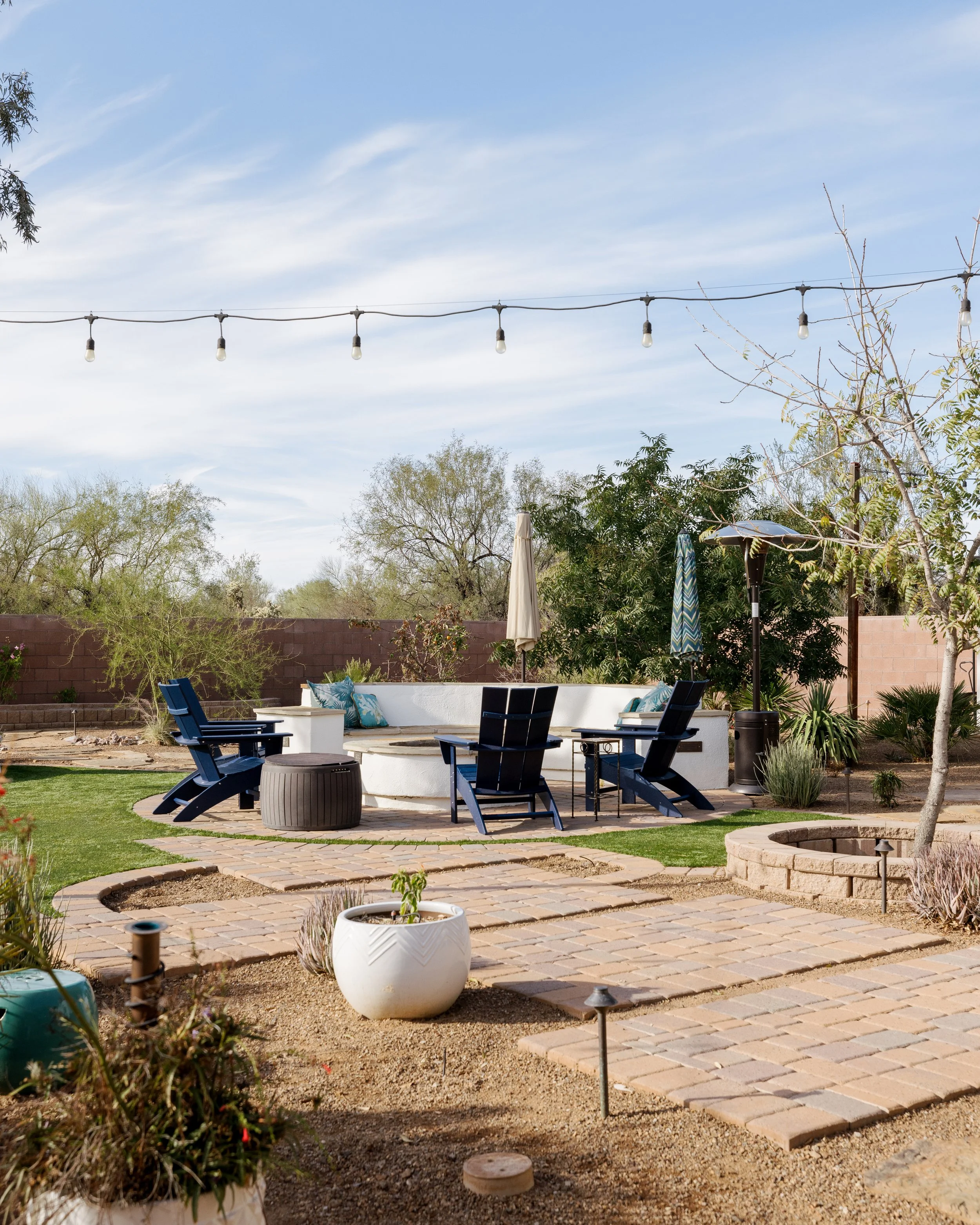 Brick walkways connect a fire pit surrounded by blue Adirondack chairs, raised planting beds, string lights, and desert landscaping in a spacious backyard.