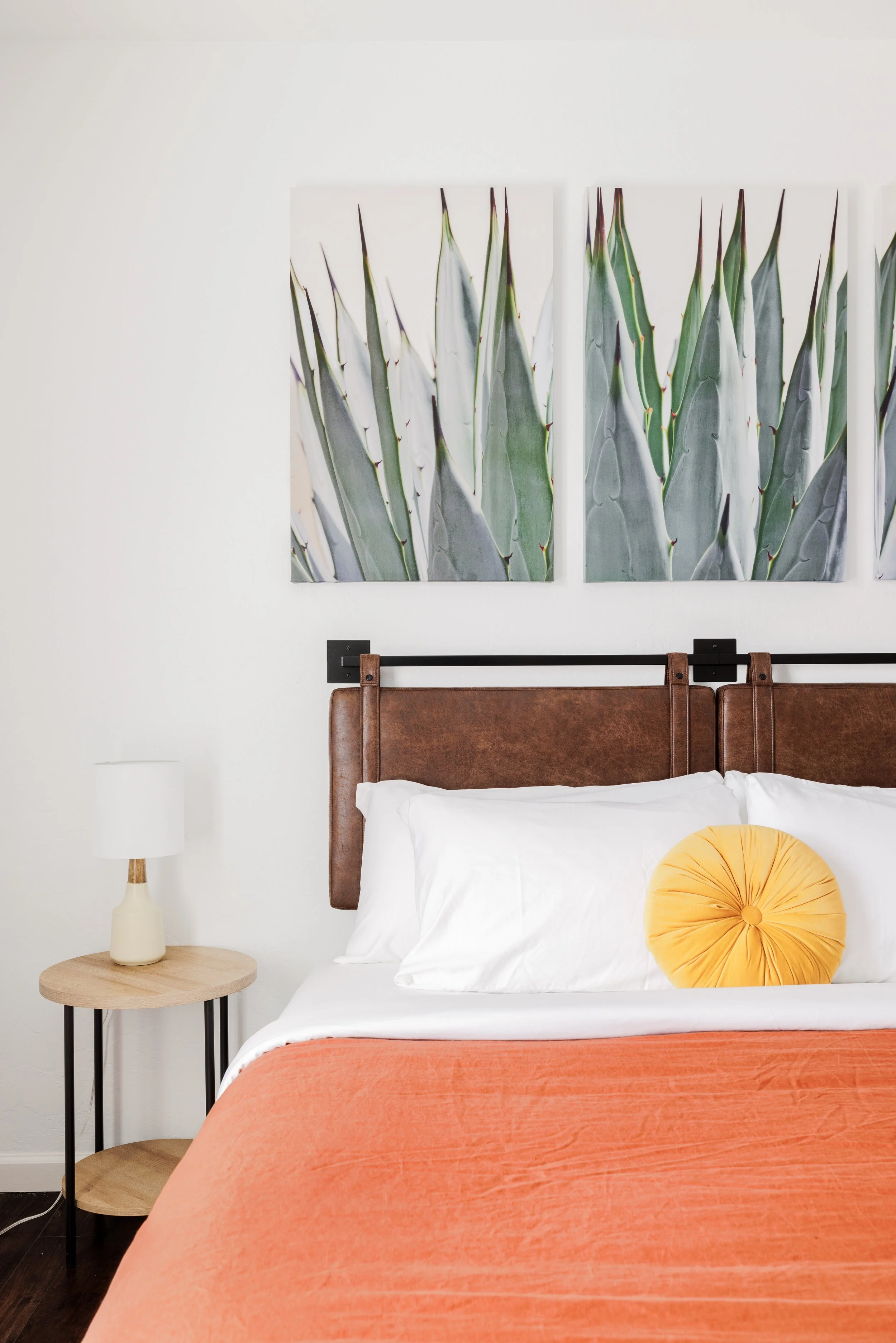 King bedroom with agave artwork above the bed, white bedding, coral duvet, leather headboard, and side table with a bedside lamp.