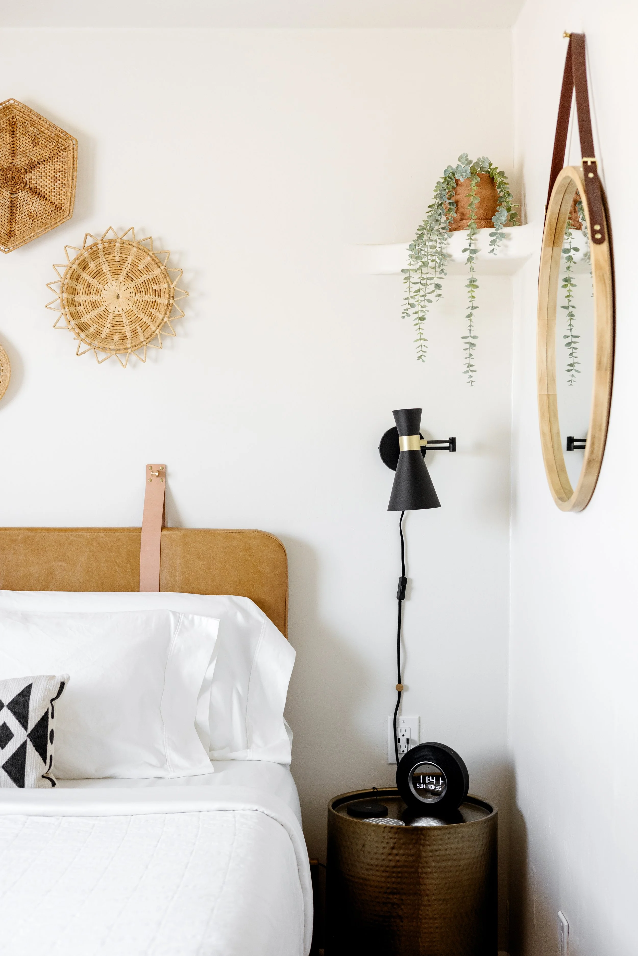 Bedroom with yellow upholstered  headboard, wall décor, round mirror, trailing plant, and black wall sconce.