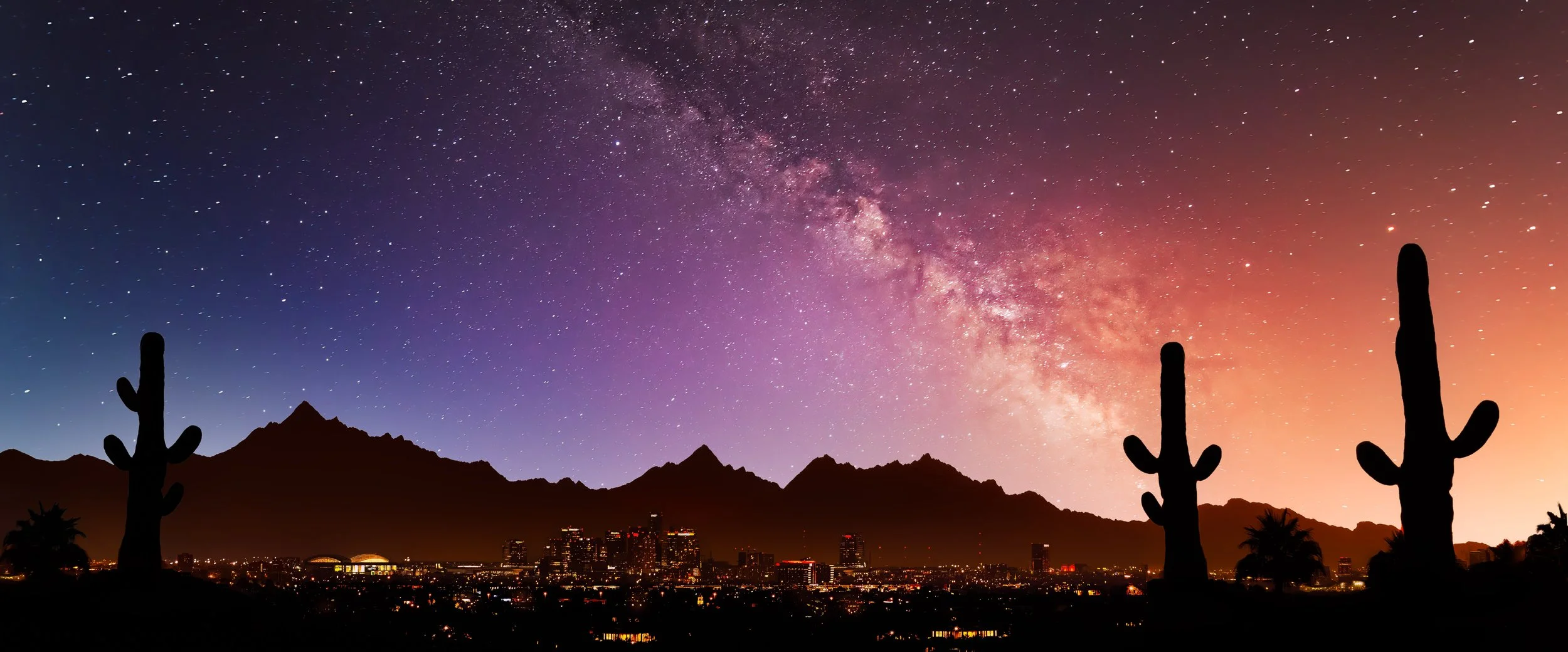 The Milky Way stretches across a colorful night sky above Tucson city lights, with dark mountain ridges and tall saguaro silhouettes in the foreground.