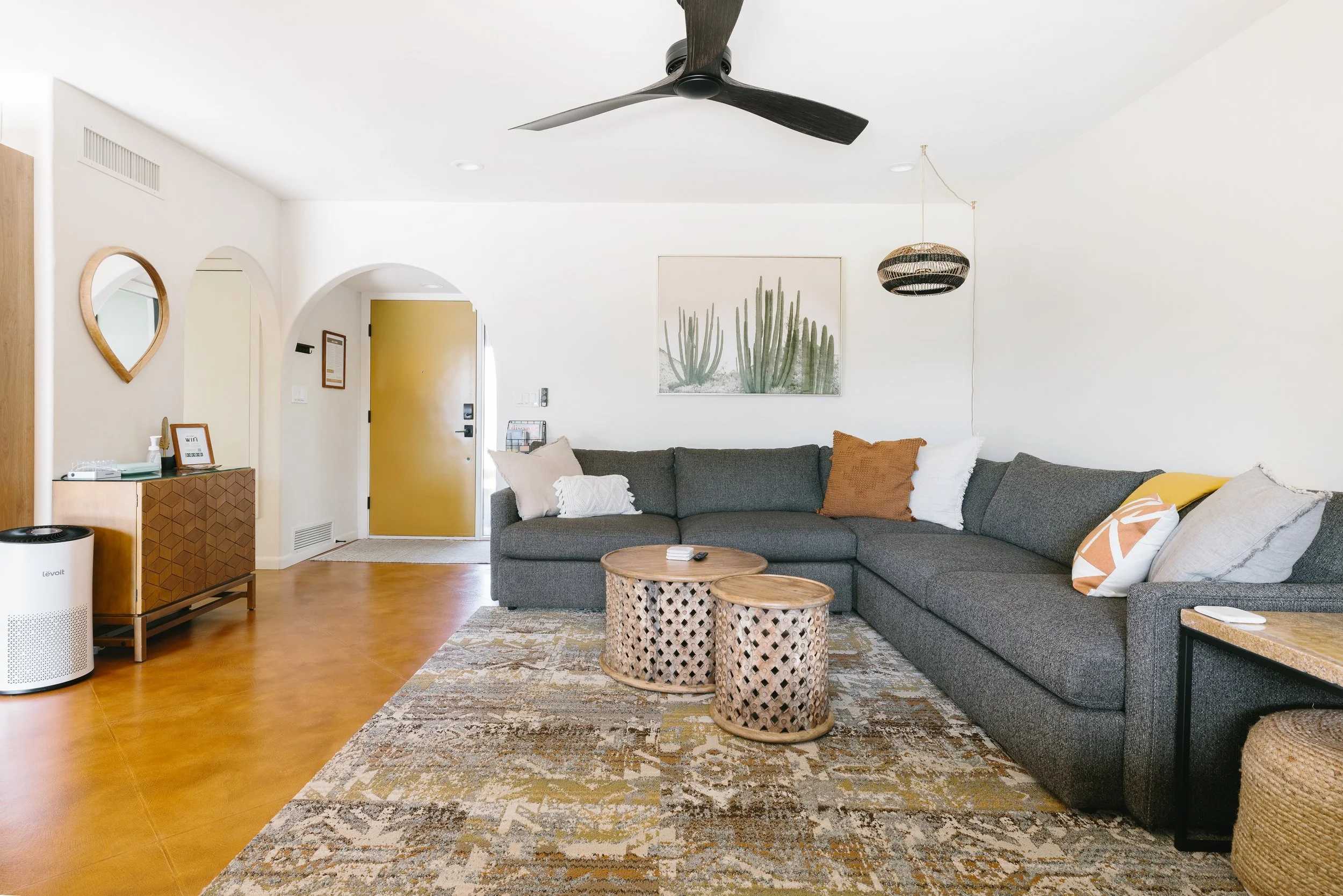 Bright living room with a large dark gray sectional sofa, patterned pillows, two round woven coffee tables, a cactus print, and a mustard yellow front door.
