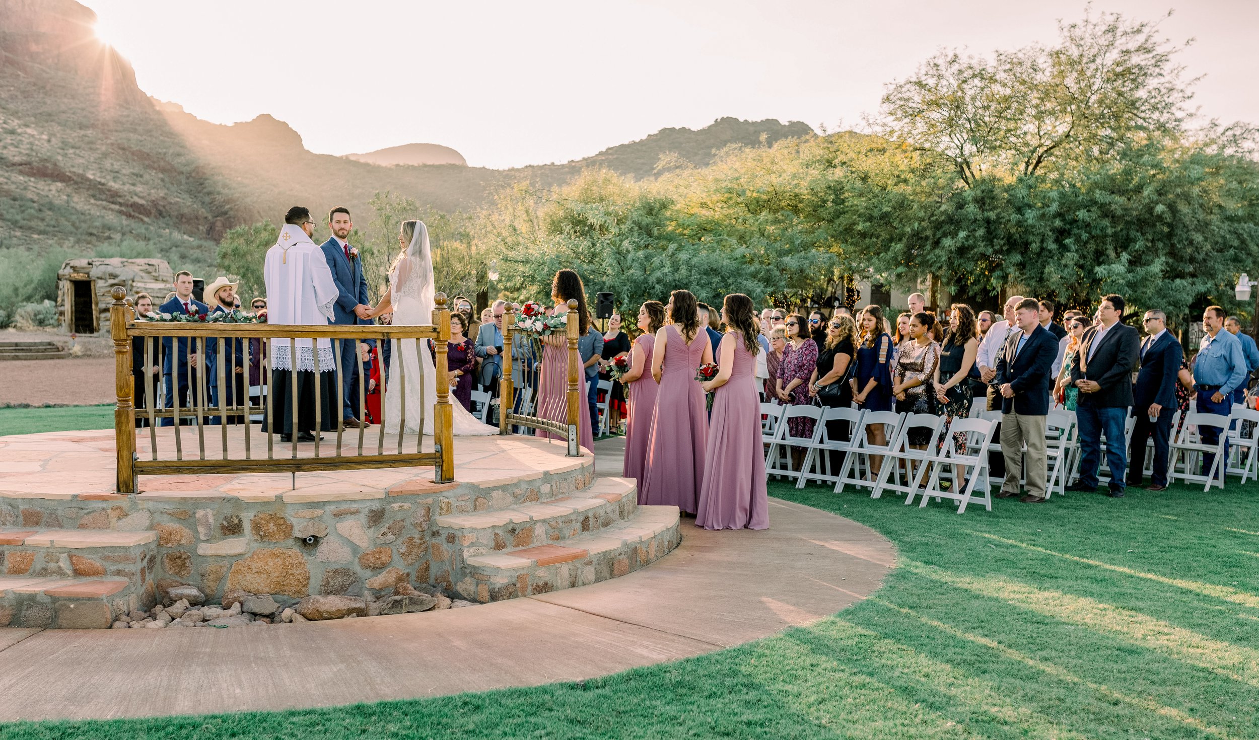 Bride and groom stand with an officiant on a raised stone platform as bridesmaids and wedding guests gather outdoors beneath desert mountains.