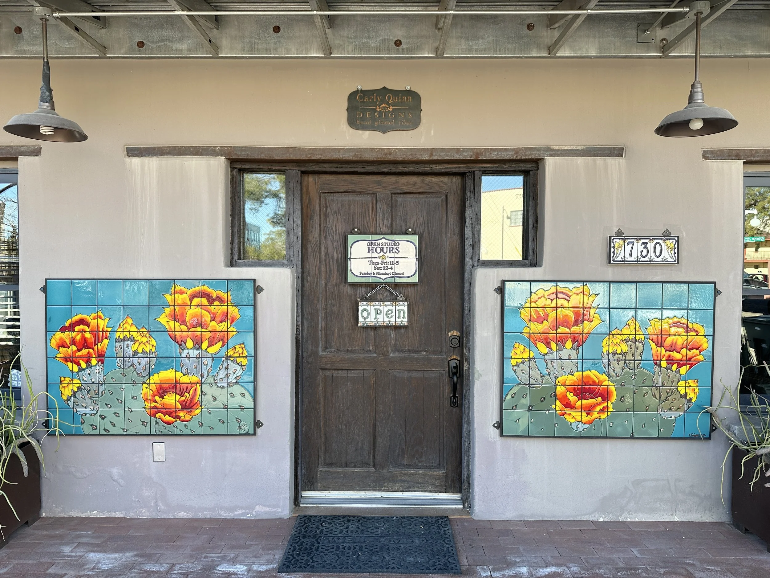 Wooden entrance to Carly Quinn Designs flanked by two large tile murals of orange cactus flowers on turquoise backgrounds and an address plaque reading 730.