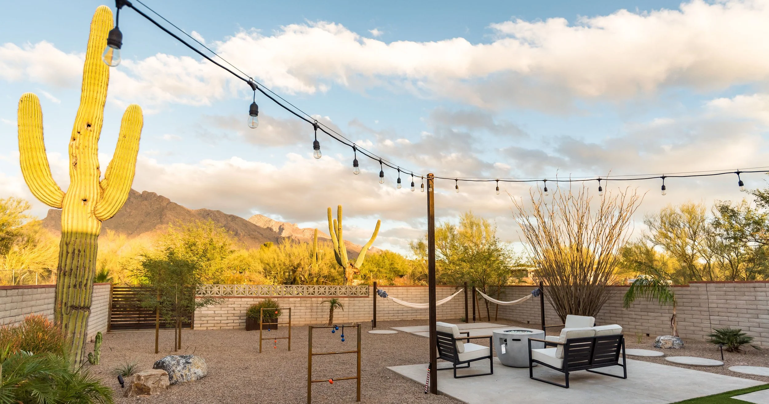 Backyard with a tall saguaro, string lights, two white hammocks, cushioned patio chairs, gravel landscaping, and mountains glowing in late-afternoon light.
