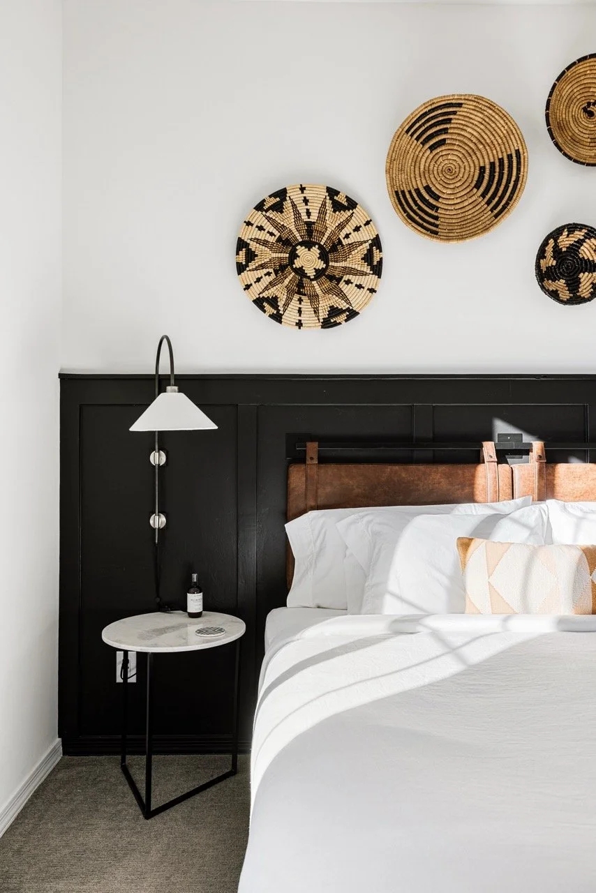 Black paneled wainscoting frames a hanging leather headboard with white bedding and tan geometric accent pillow. Three decorative woven baskets hang above the bed, and a round white marble nightstand sits beneath a wall-mounted reading lamp.