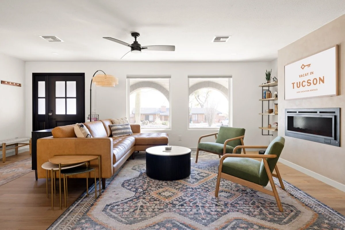 Living room with a caramel leather sectional, two green armchairs, patterned rug, round coffee table, fireplace, and open wall shelving.