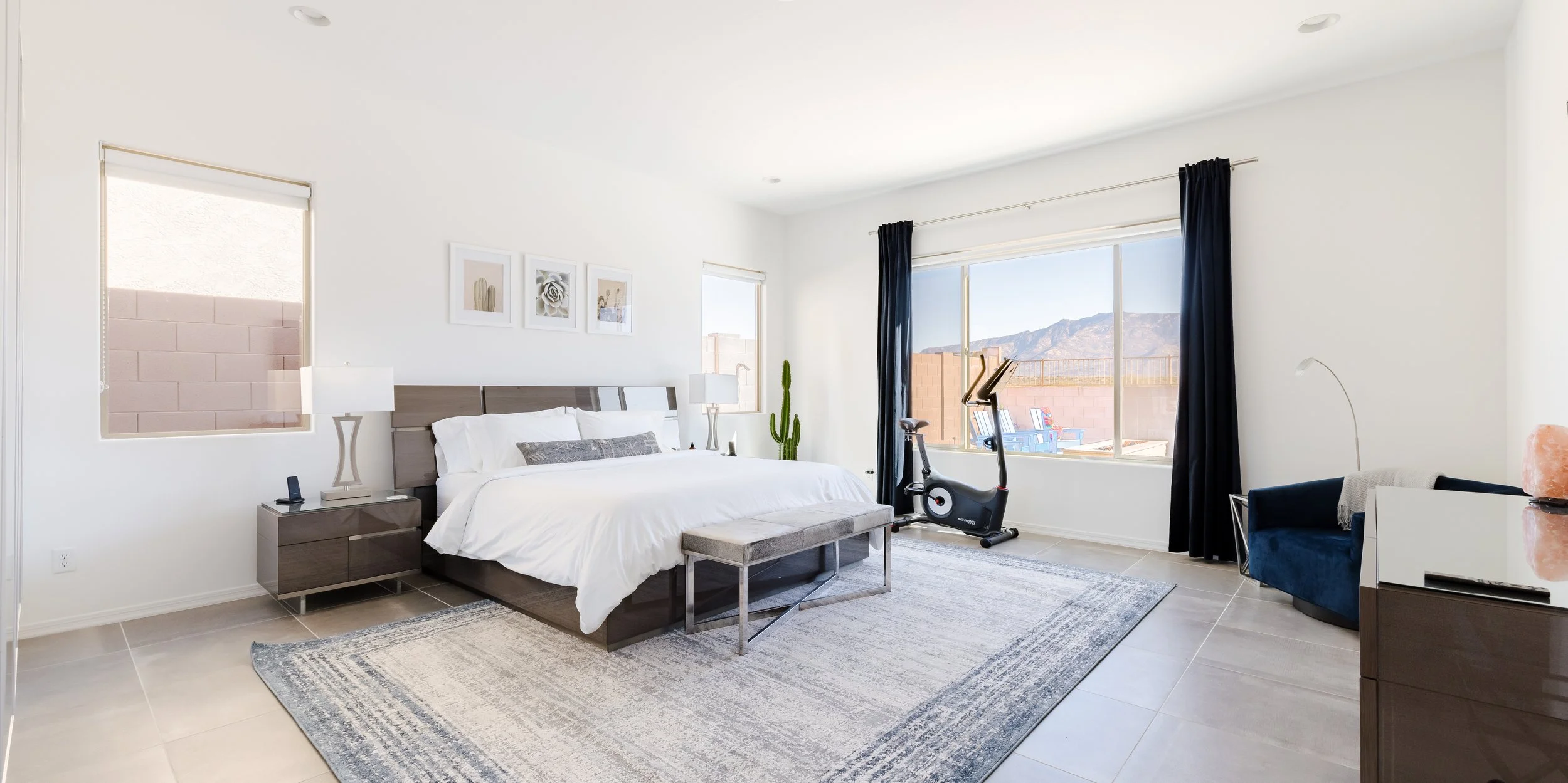 Large bright bedroom with a white bed, gray upholstered bench, blue accent chair, exercise bike, dark curtains, area rug, and mountain views through the window.