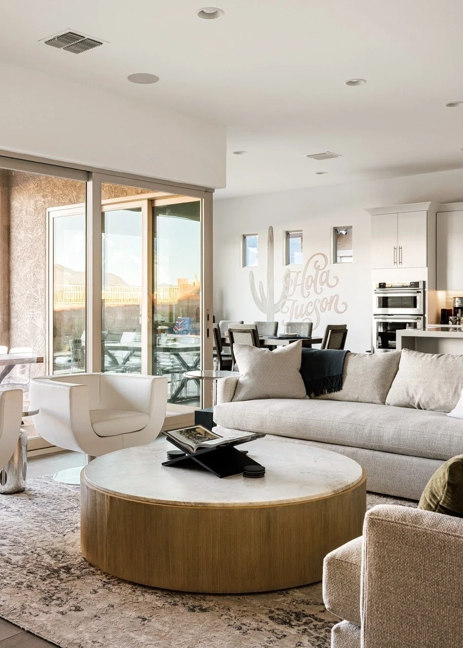 Bright open living room with white sofas, round wood coffee table, white accent chairs, and large sliding glass doors opening to the patio. The kitchen, dining room, and mural which reads "Hola Tucson" can be seen in the background.