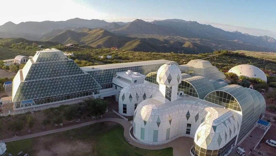 Aerial view of the Biosphere 2 complex with large glass pyramids, curved greenhouse structures, white domed buildings, and desert mountains in the background.