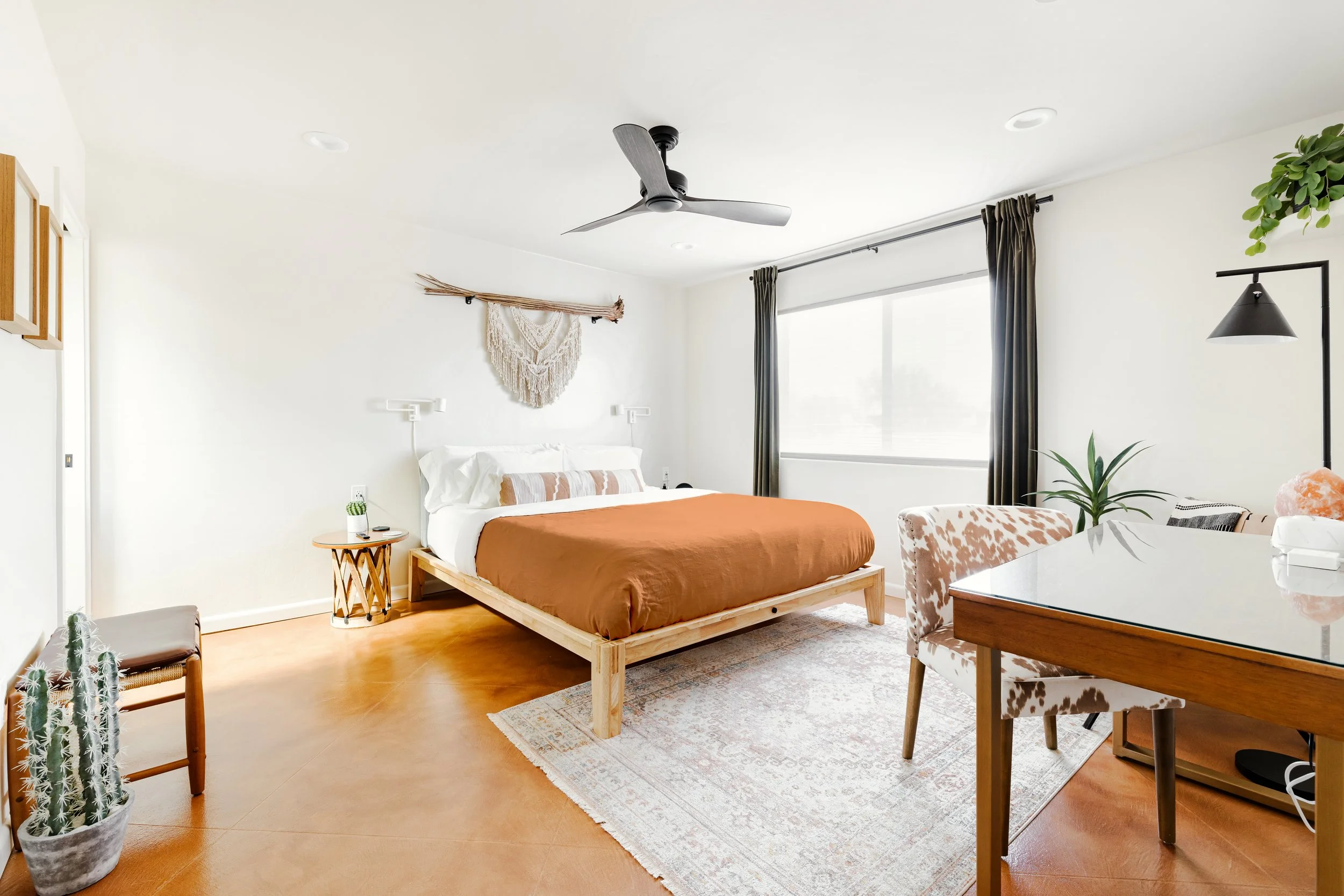Bright bedroom with a large wood-frame bed, rust-colored duvet, white bedding, cowhide-patterned chair, writing desk, cactus plants, and macramé wall hanging.