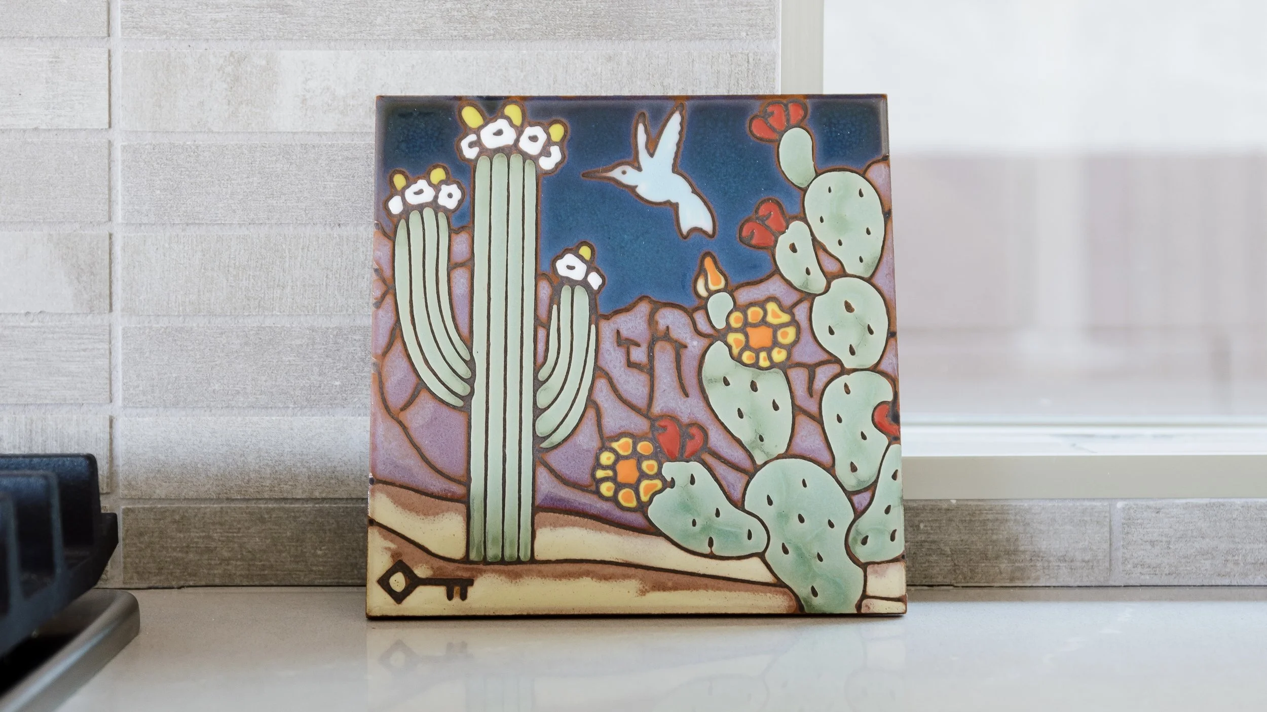 Square ceramic tile artwork showing a saguaro cactus, prickly pear, hummingbird, desert mountains, flowers, and a small key motif against a deep blue background.