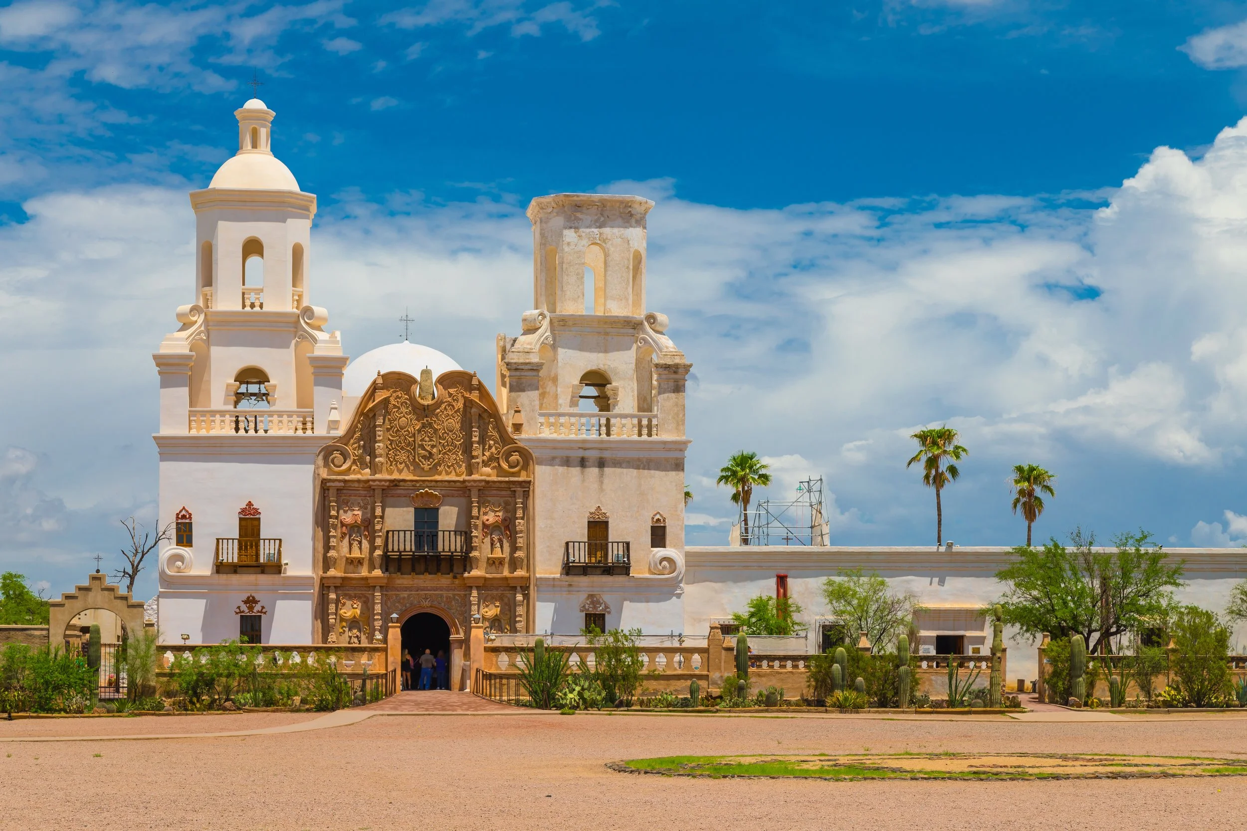 Historic white church with twin bell towers, an ornate carved central facade, desert landscaping, and palm trees beneath a blue sky with large white clouds.