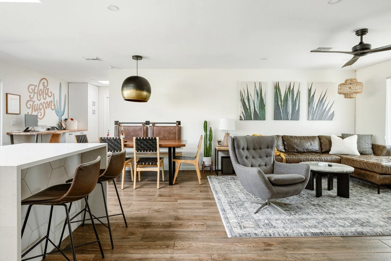 Warm wood floors connect the dining and living areas, where leather dining chairs surround a wood table.  A gray armchair, cactus artwork, and a large black and gold pendant light create a clean mid-century desert style.
