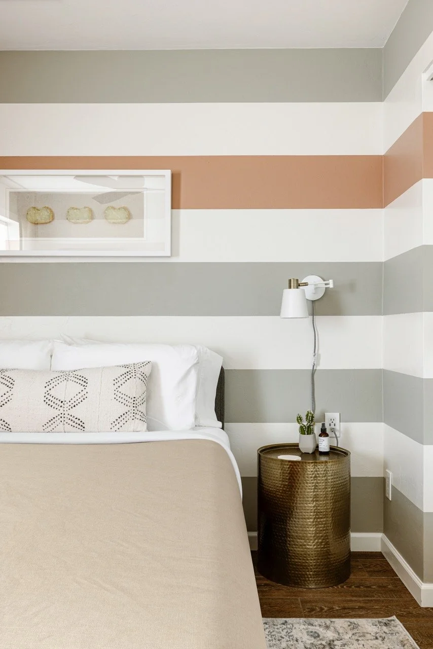 A neutral linen bedspread sits in front of walls painted with wide horizontal stripes in muted desert tones. Framed agate artwork hangs above the bed, with a brass barrel nightstand, wall-mounted light to the right.