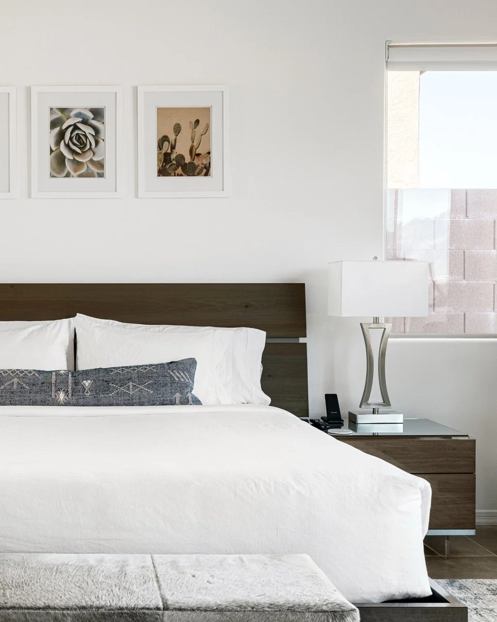 King bed with crisp white bedding sits against a dark wood headboard. A modern lamp sits on the bedside table to the right, and framed desert botanical artwork hangs above, while a light upholstered bench sits at the foot of the bed.