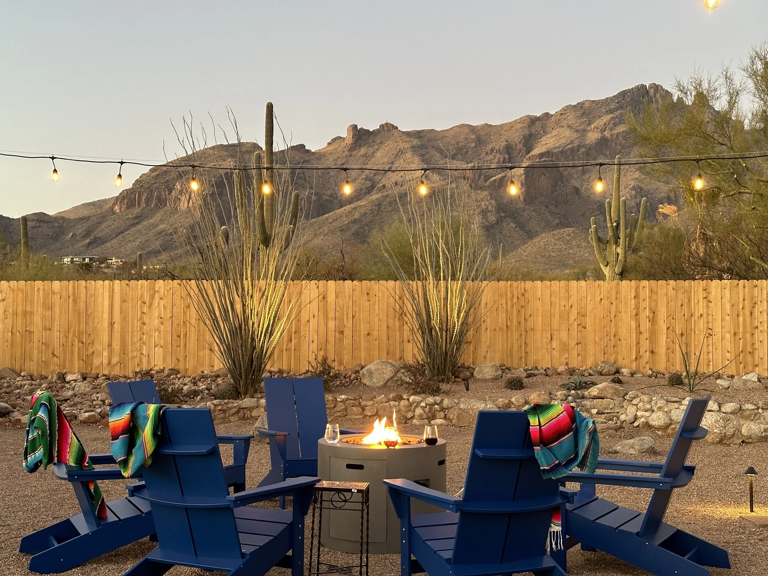 Blue Adirondack chairs surround a fire pit with desert plants and mountain views in the background.