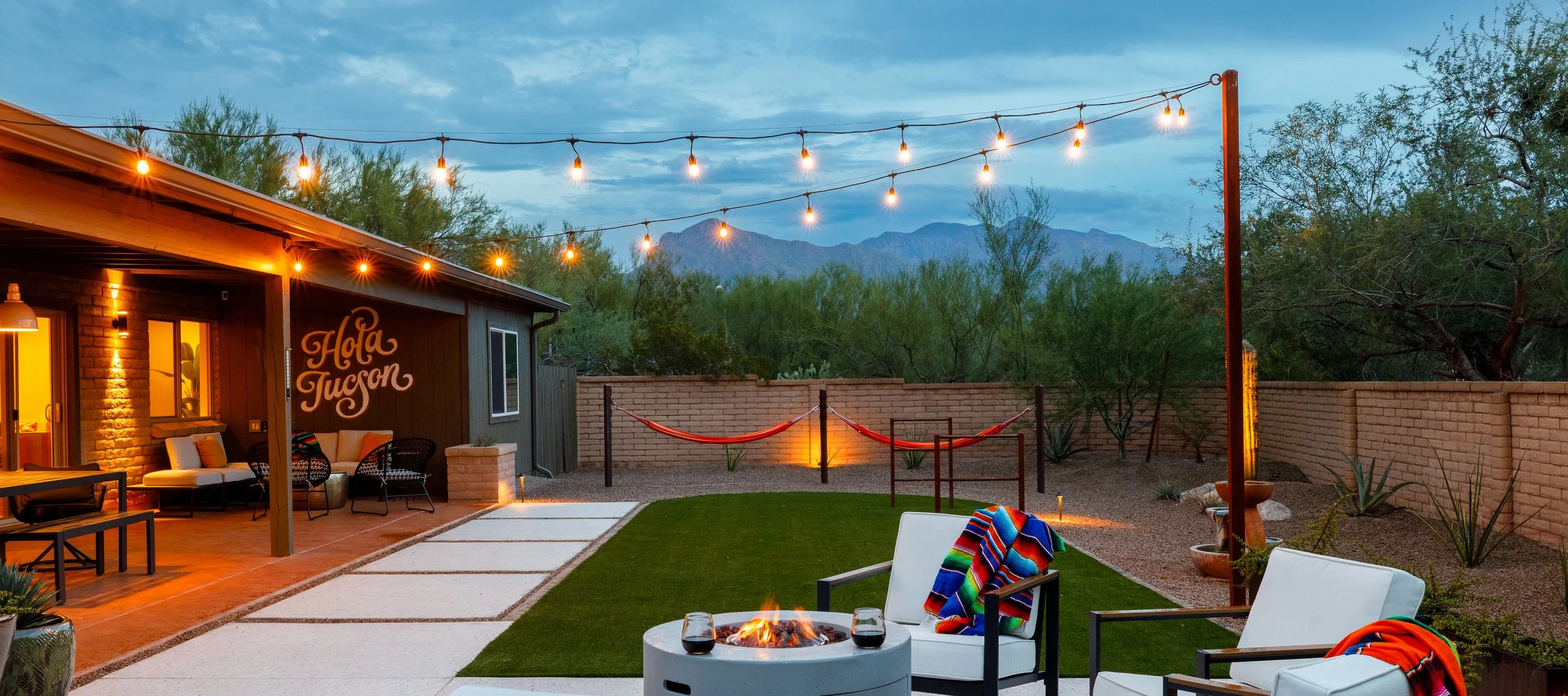 Backyard at dusk with glowing string lights, a round fire pit surrounded by cushioned chairs, two hammocks, green turf, desert plants, and mountains in the distance.
