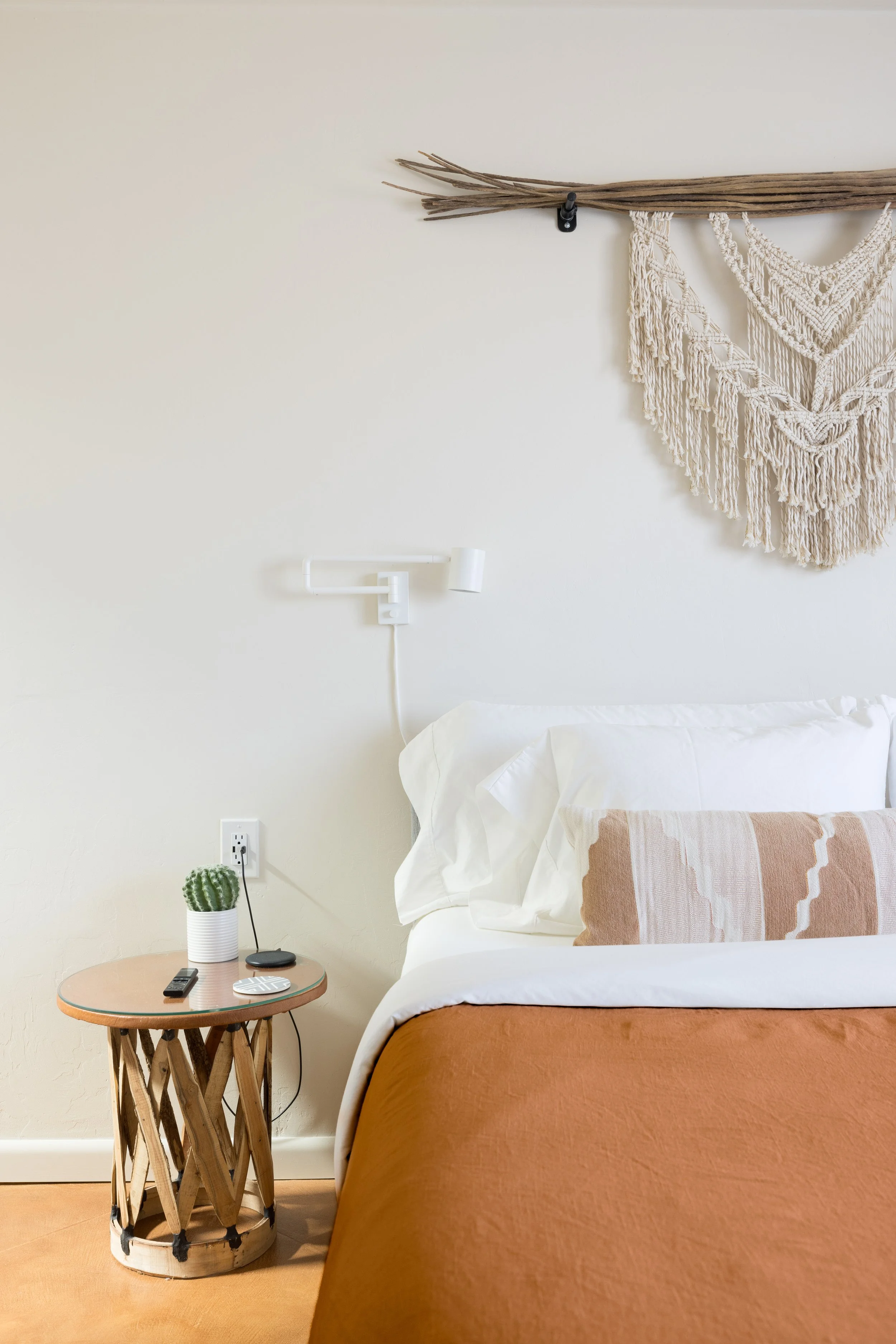 Bedroom with a California king bed, macramé wall hanging, round wood side table, and rust-colored duvet and white bedding.