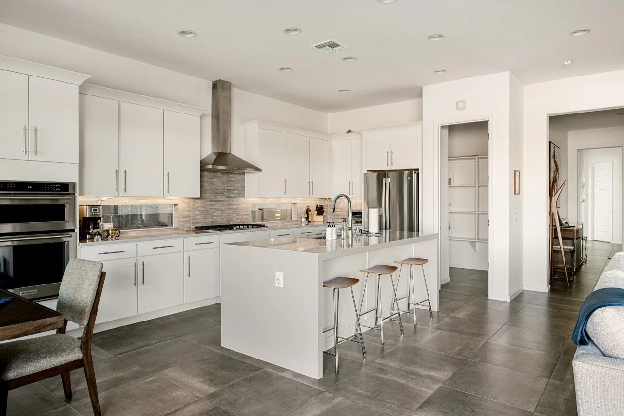 Large white kitchen with a waterfall island, three backless bar stools, stainless steel appliances, gray tile backsplash, vent hood, and wide gray floor tile. A walk-in pantry can be seen in the right corner.