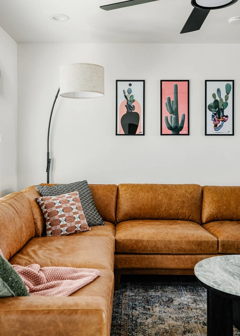 Open-concept living room with a caramel leather sectional, round coffee table, desert-inspired artwork, and large windows overlooking the Catalina Mountains in this Tucson vacation rental.