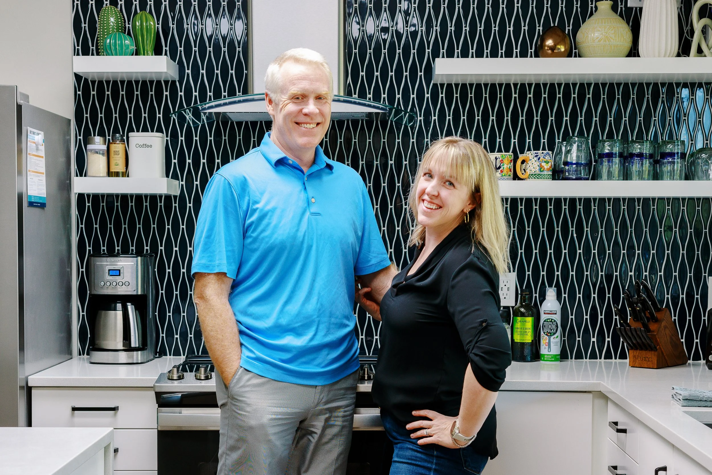 Gray-haired man in a blue polo and blonde woman in a black blouse smile together in a modern kitchen with a black geometric tile backsplash.