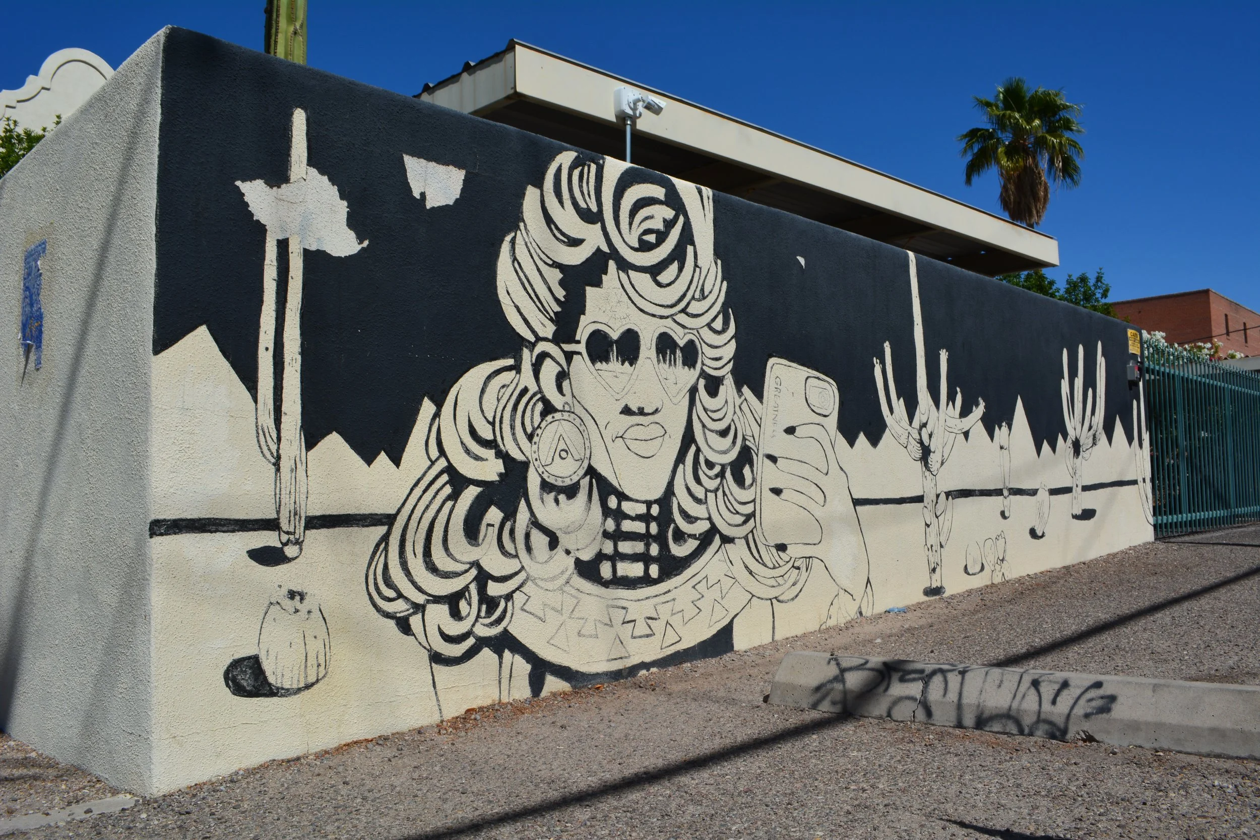 Black-and-white mural of a woman with heart-shaped sunglasses holding a phone, surrounded by saguaros and desert silhouettes.