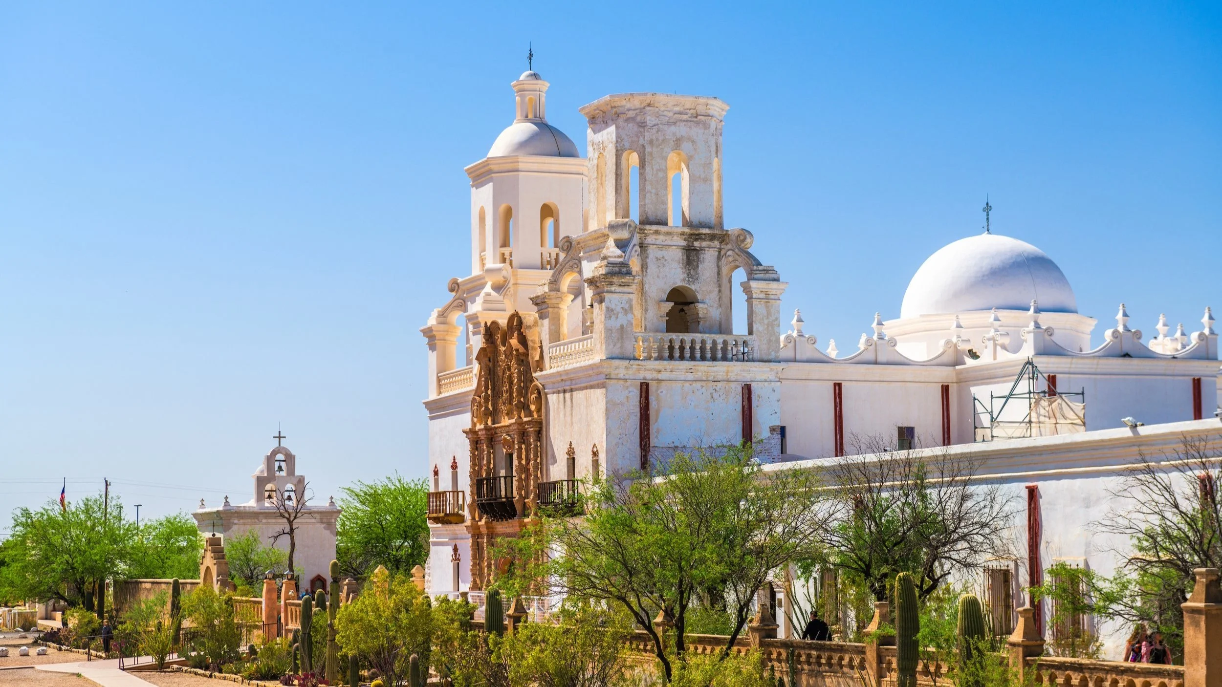 Historic white mission with ornate towers and domes rises above desert landscaping under a clear blue sky.