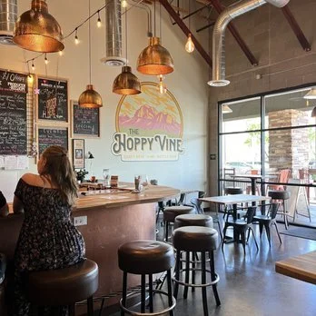 Interior of The Hoppy Vine in Oro Valley with a woman seated at the bar, copper pendant lights, high-top seating, chalkboard menus, and the Hoppy Vine logo on the wall.