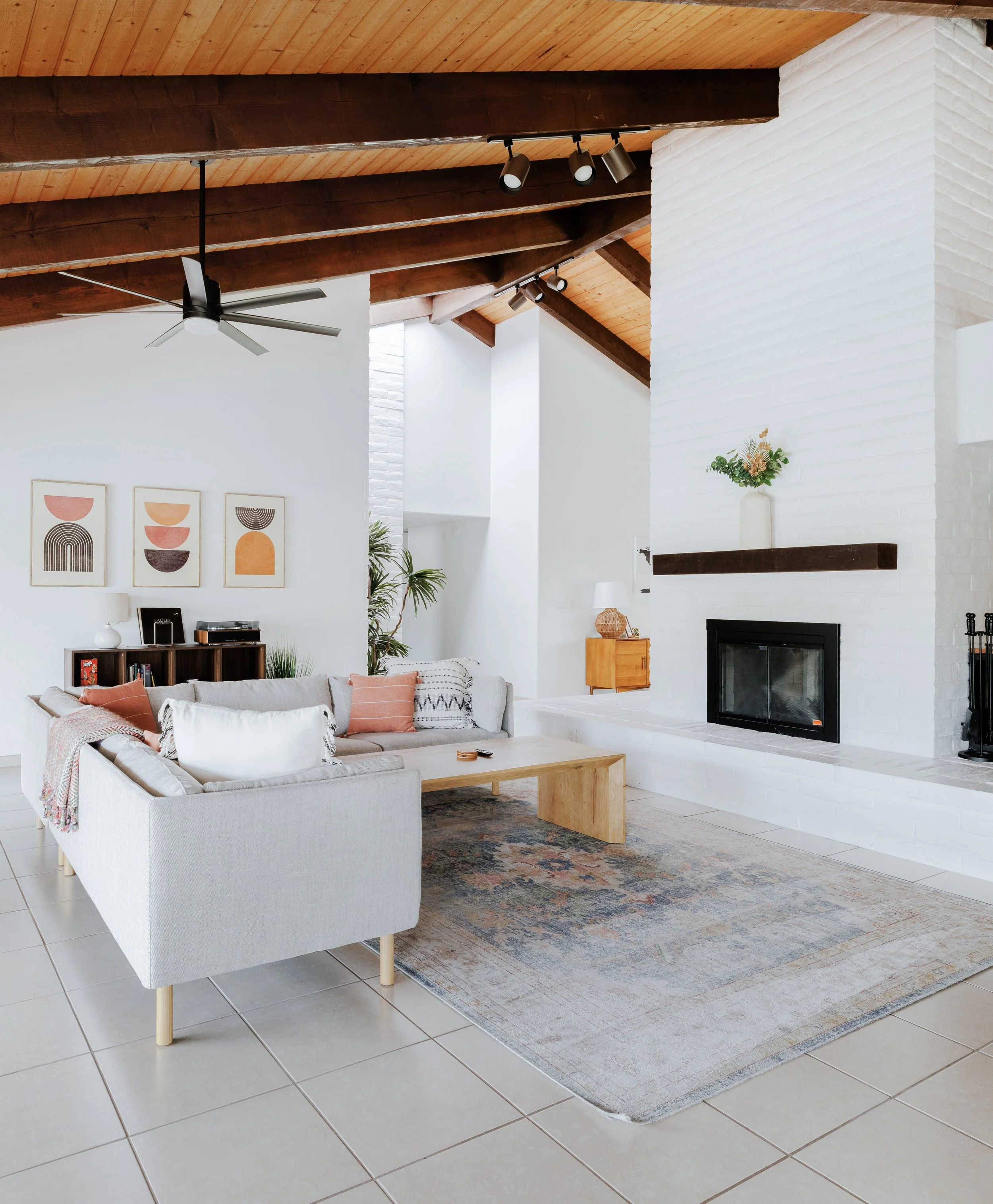 Open living room with vaulted wood ceilings, a dual-sided fireplace, white corner sofa with wooden coffee table, a record player, and a large area rug.