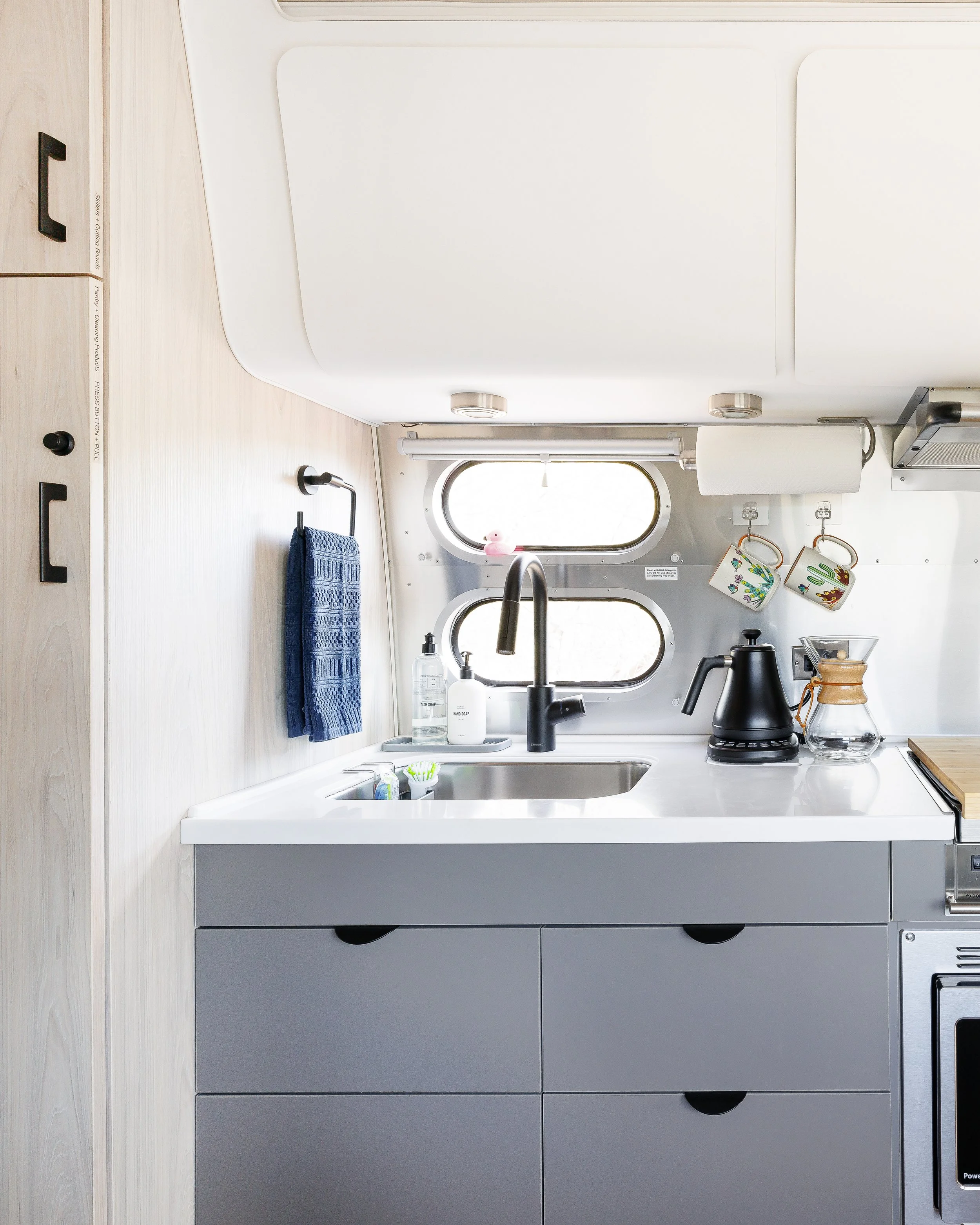 Compact kitchen with a sink, lower cabinets, countertop appliances, hanging towels, and windows above the sink.