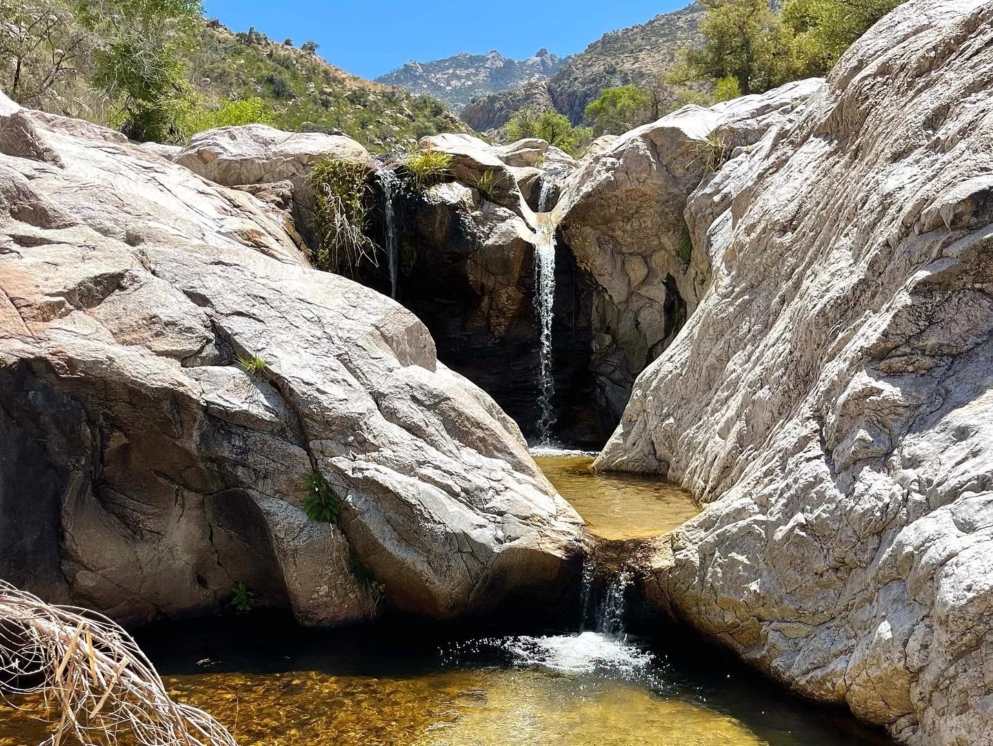 Small waterfalls cascade over large granite boulders into clear desert pools surrounded by rugged mountains.