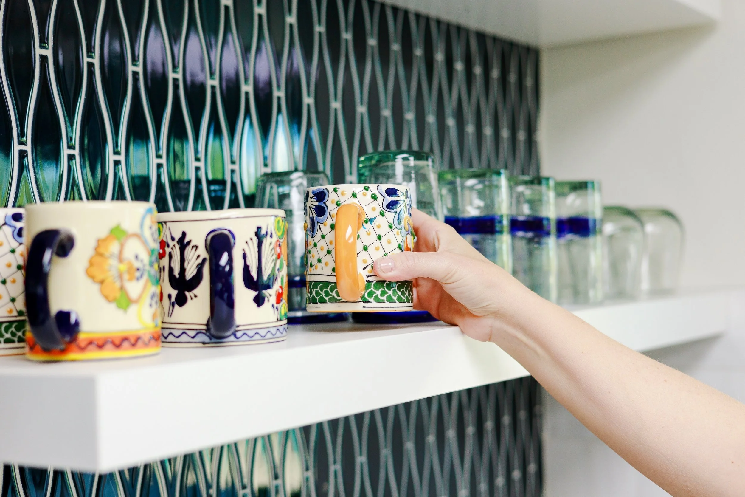 Hand reaches for a colorful hand-painted ceramic mug displayed on an open kitchen shelf against a black geometric tile backsplash.